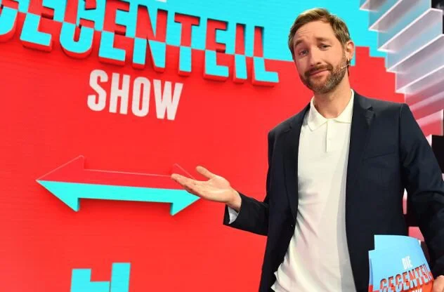 A man in a dark suit and white shirt stands in front of a red background with the poster 'The Great American Congregational Show'. He is gesturing with his hand towards the left with a slight smile.