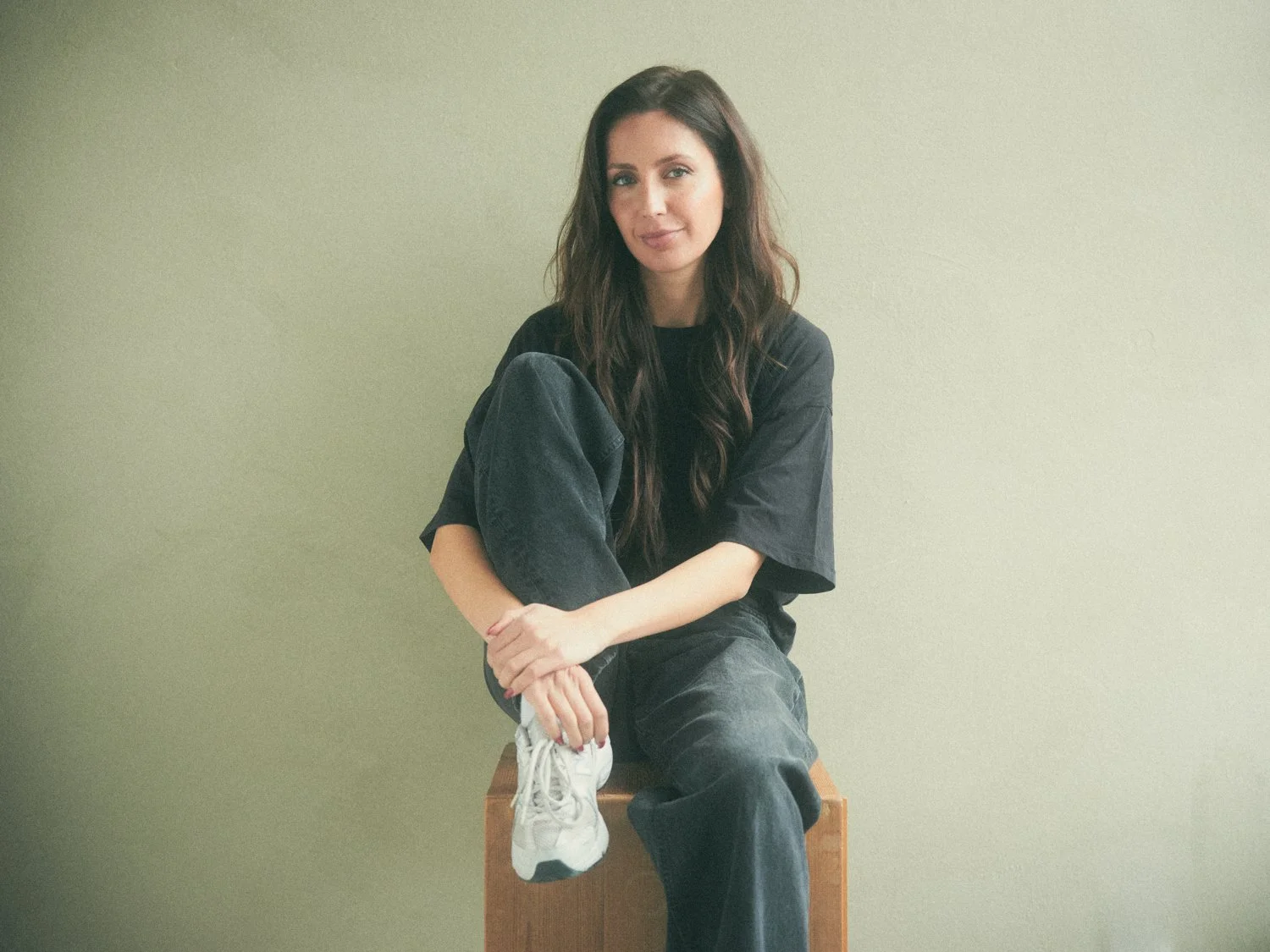 A woman with long dark hair sits on a wooden block, wearing a black t-shirt, black jeans, and white sneakers, against a plain light green wall.