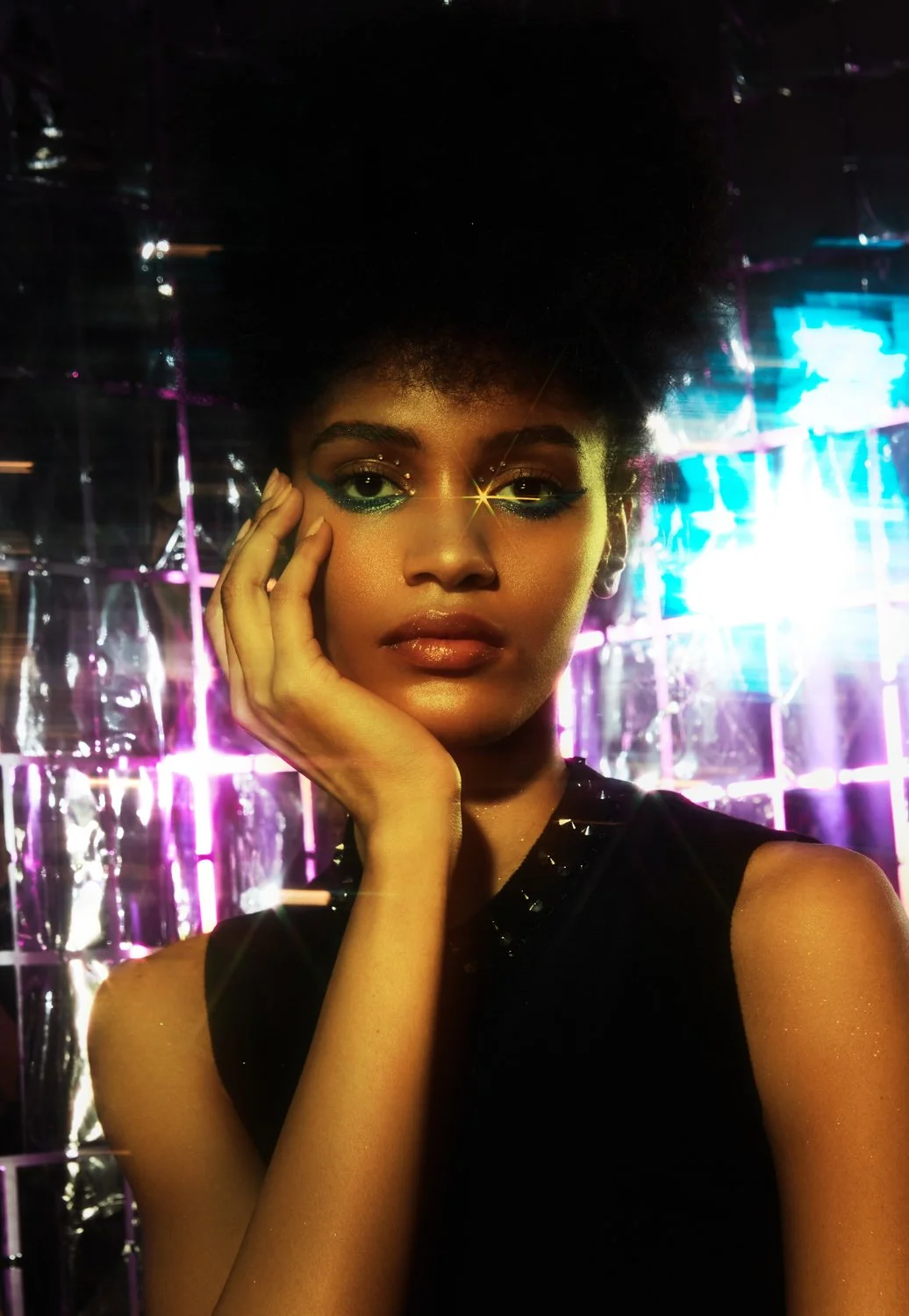 Young woman with bold makeup posing in front of a shiny, colorful backdrop.