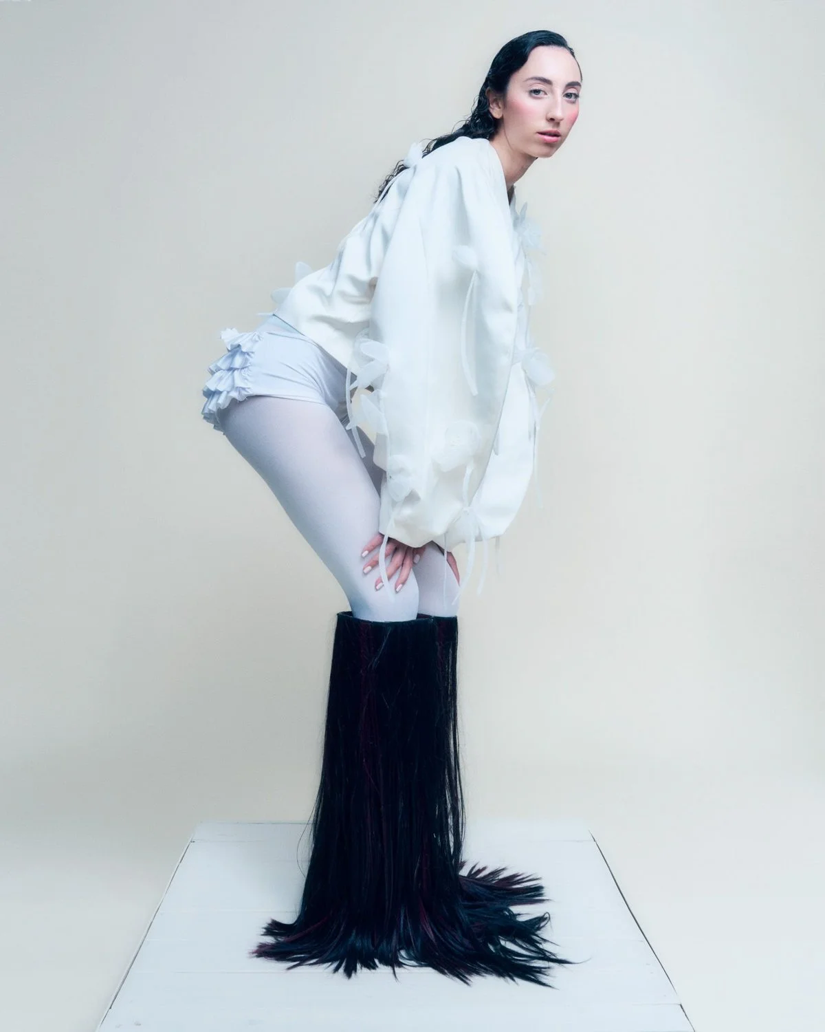 A woman with dark hair posing against a plain background, wearing a white top, white shorts, white tights, and black boots with long black fur extending from the boots.