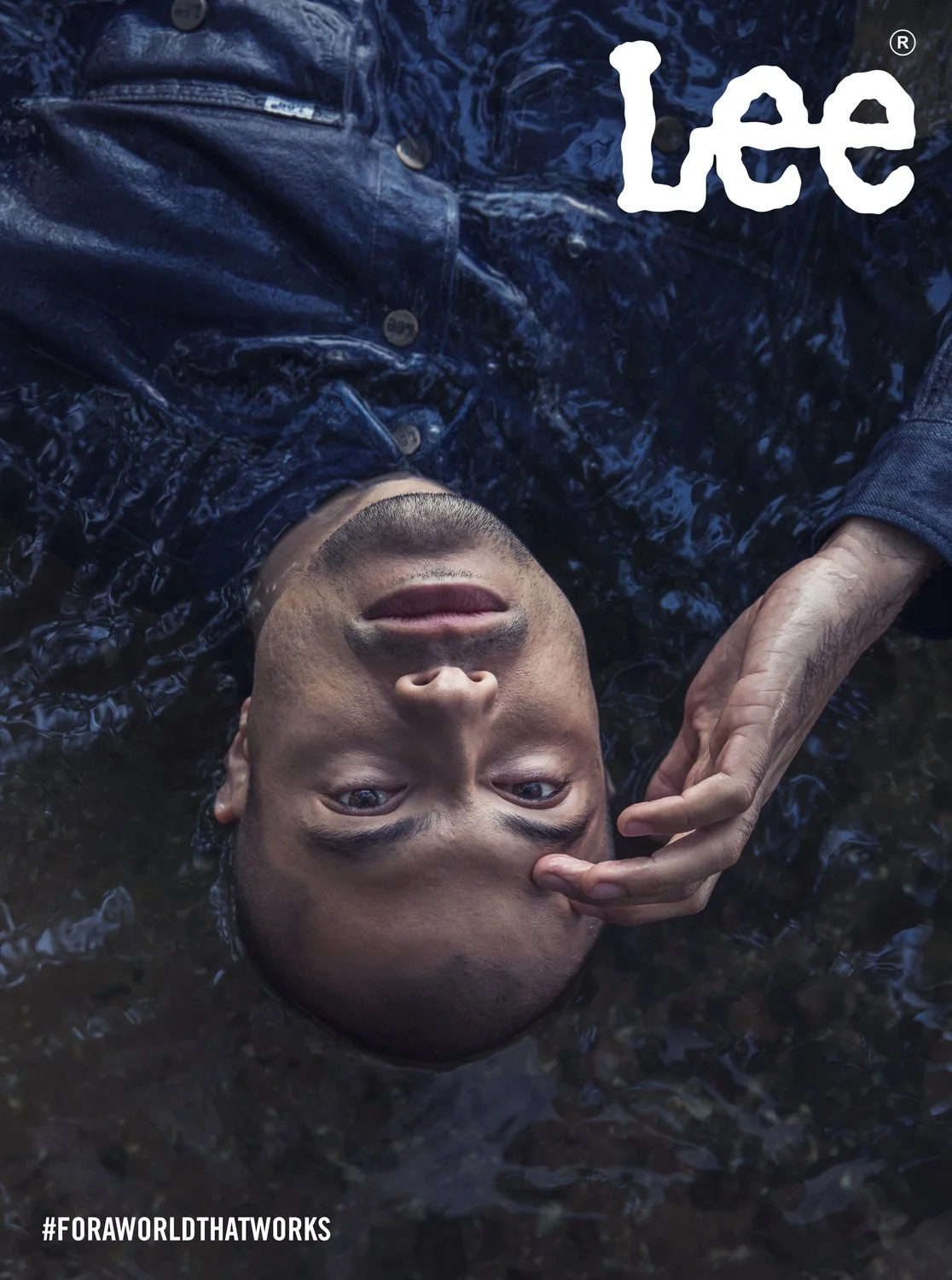 A man lies in water, wearing a dark shiny jacket, with one hand near his face. The photo is taken from above, and the word 'Lee' appears in the upper right corner. The hashtag '#FORAWORLTHATWORKS' is at the bottom left.