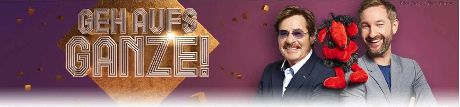 Two men smiling, one with glasses and a mustache, and another with a beard, with a plush dog on his shoulder, against a purple background with a sign that says "Geh Aufs Ganze!" and gold confetti.