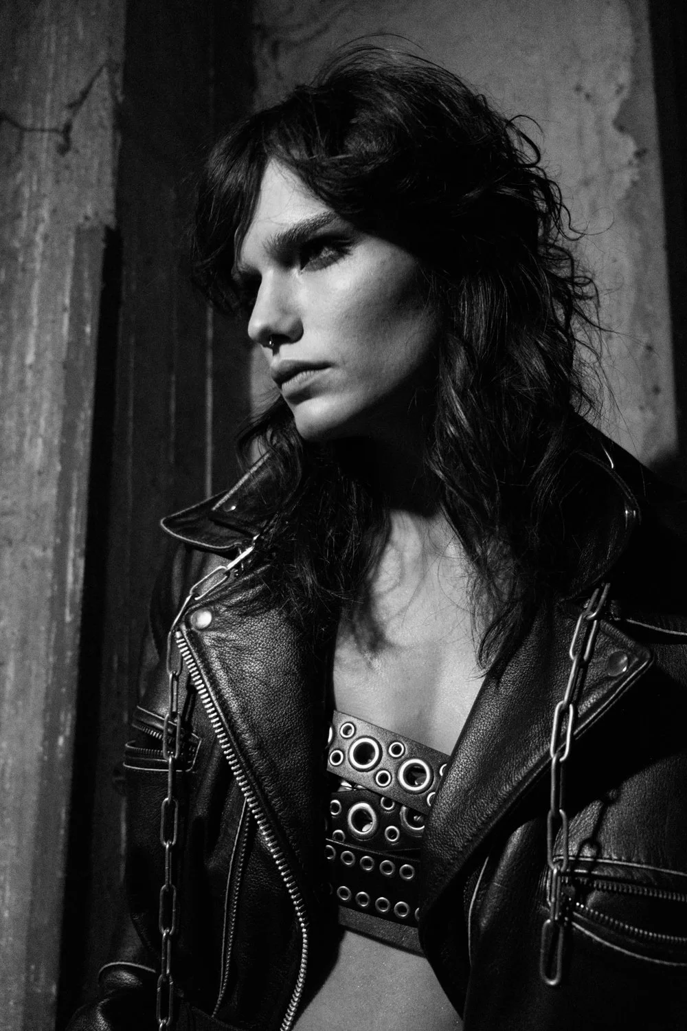 Black and white portrait of a woman with dark, wavy hair wearing a leather jacket and a studded top, standing in front of a textured wall.