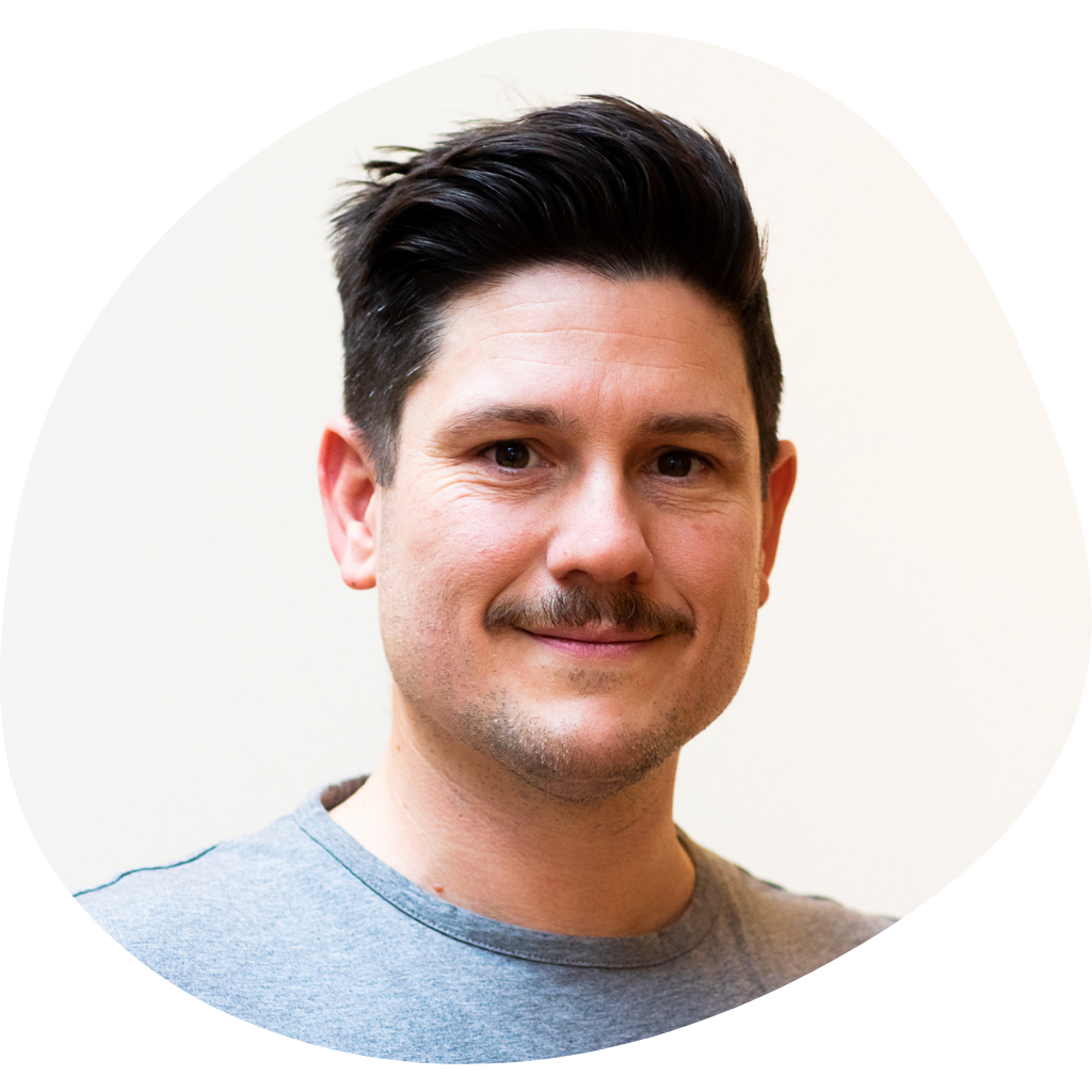 A smiling man with dark hair and a mustache, wearing a gray shirt, facing the camera against a plain light background.