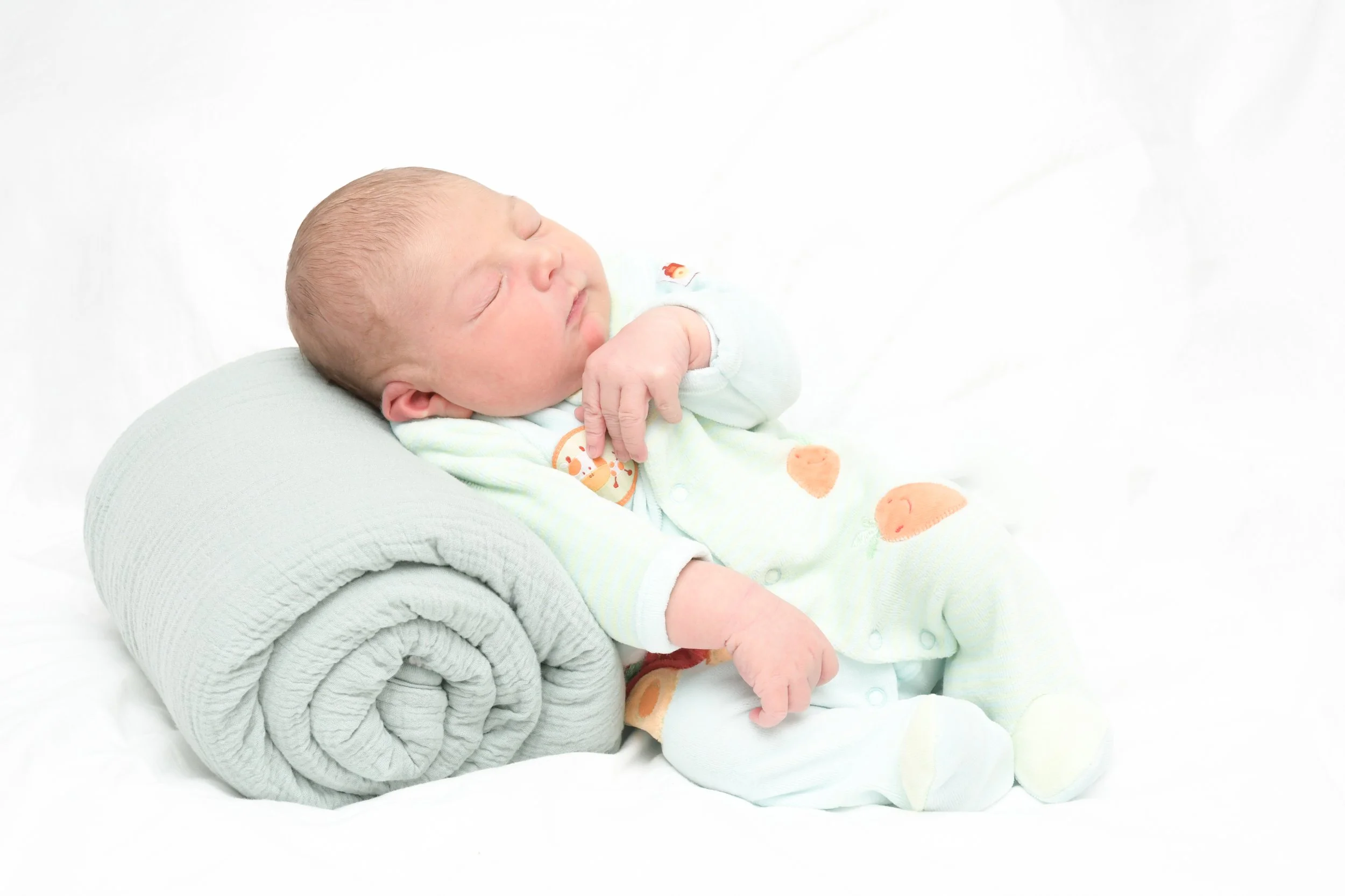 Un bébé allongé sur un coussin gris, dormant paisiblement. Le bébé porte un pyjama pastel avec des motifs d'animaux et est entouré d'un fond blanc.