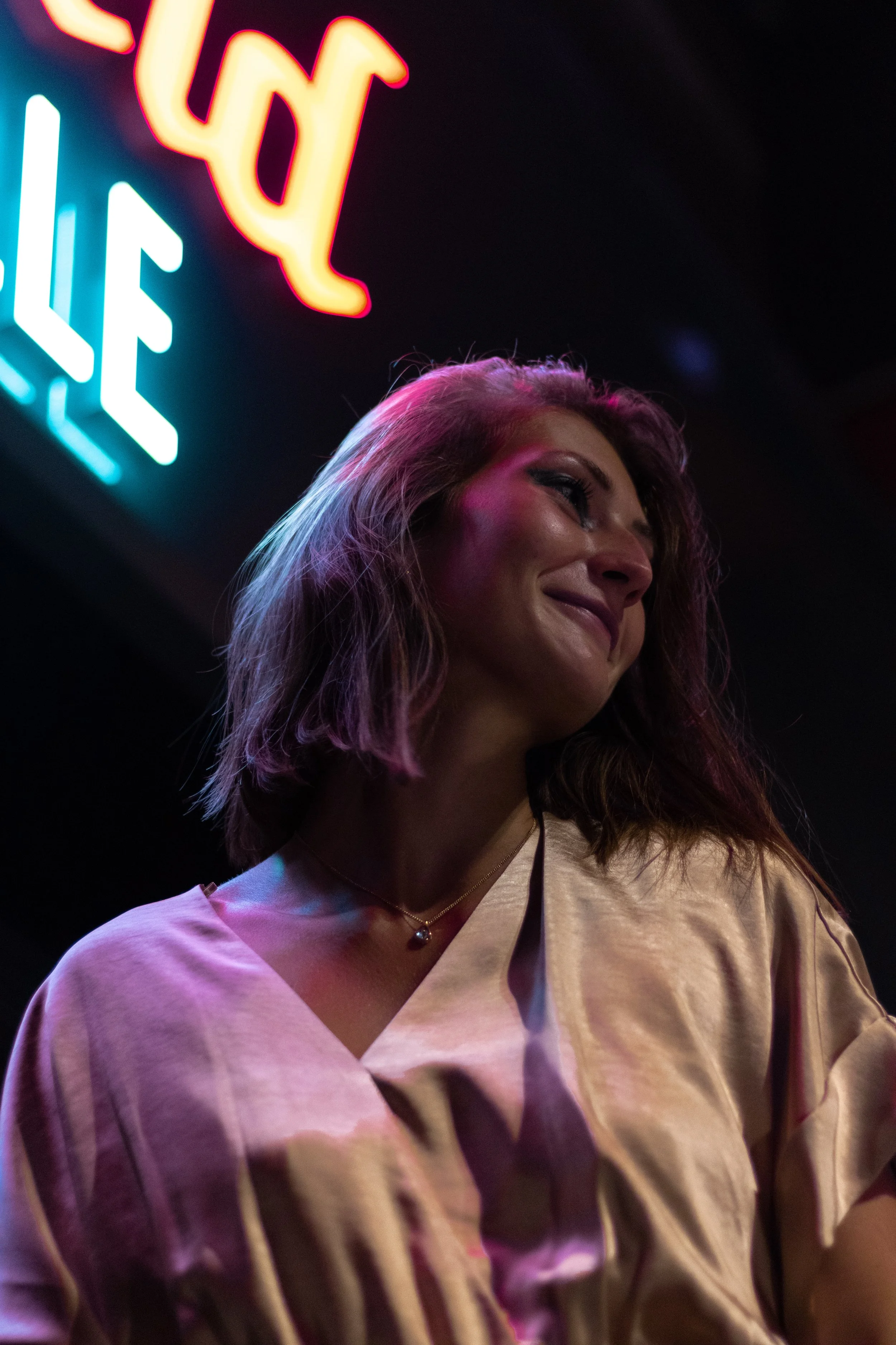 Une femme souriante avec des cheveux bruns, portant une robe satinée beige, sous un éclairage colored néon dans un environnement intérieur.