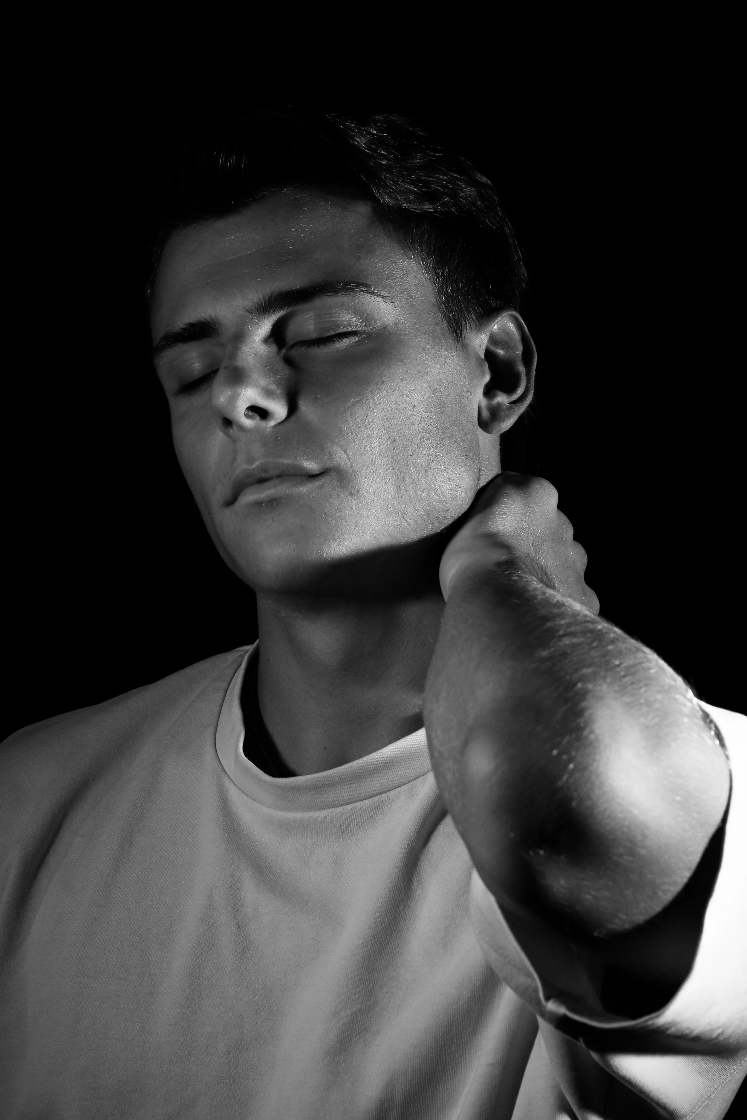 Jeune homme en noir et blanc, yeux fermés, visage détendu, se tenant le cou avec la main, portant un t-shirt clair, fond sombre.