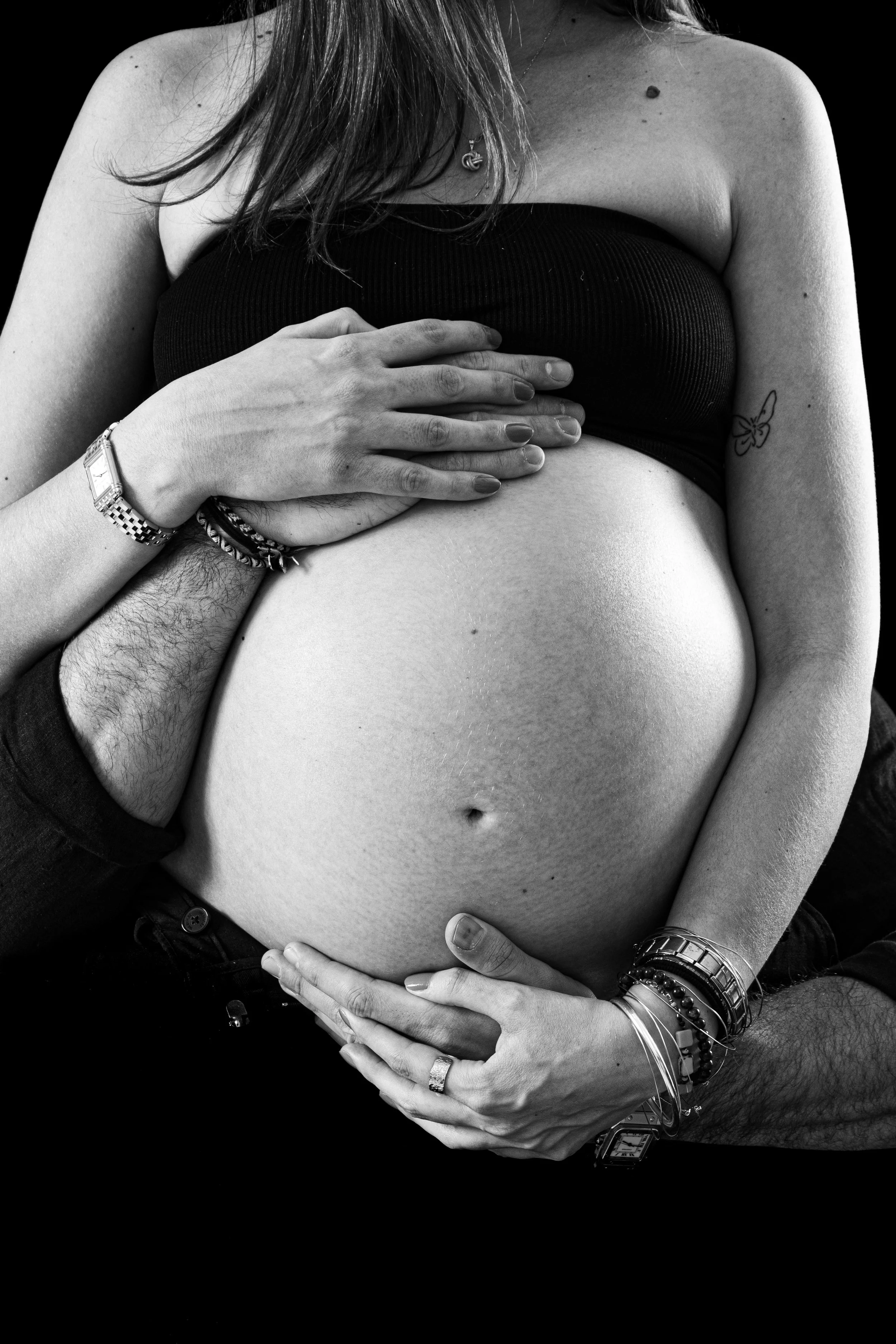 Une femme enceinte portant un haut noir, étant caressée par deux mains masculines, avec la femme portant plusieurs bracelets et une chaîne en collier, dans une ambiance en noir et blanc.