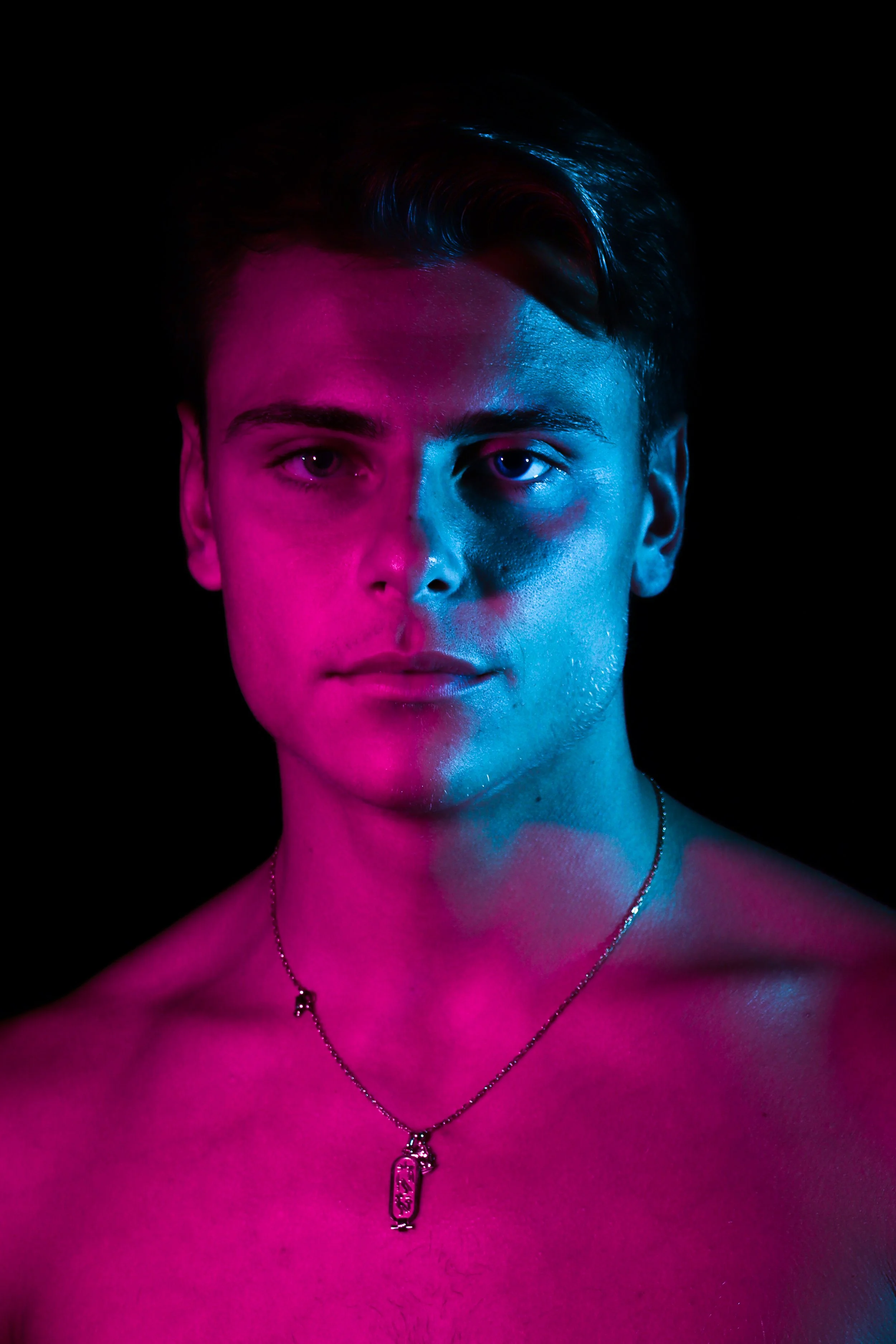 Portrait d'un jeune homme nu, éclairé par des lumières bleues et roses sur fond noir.