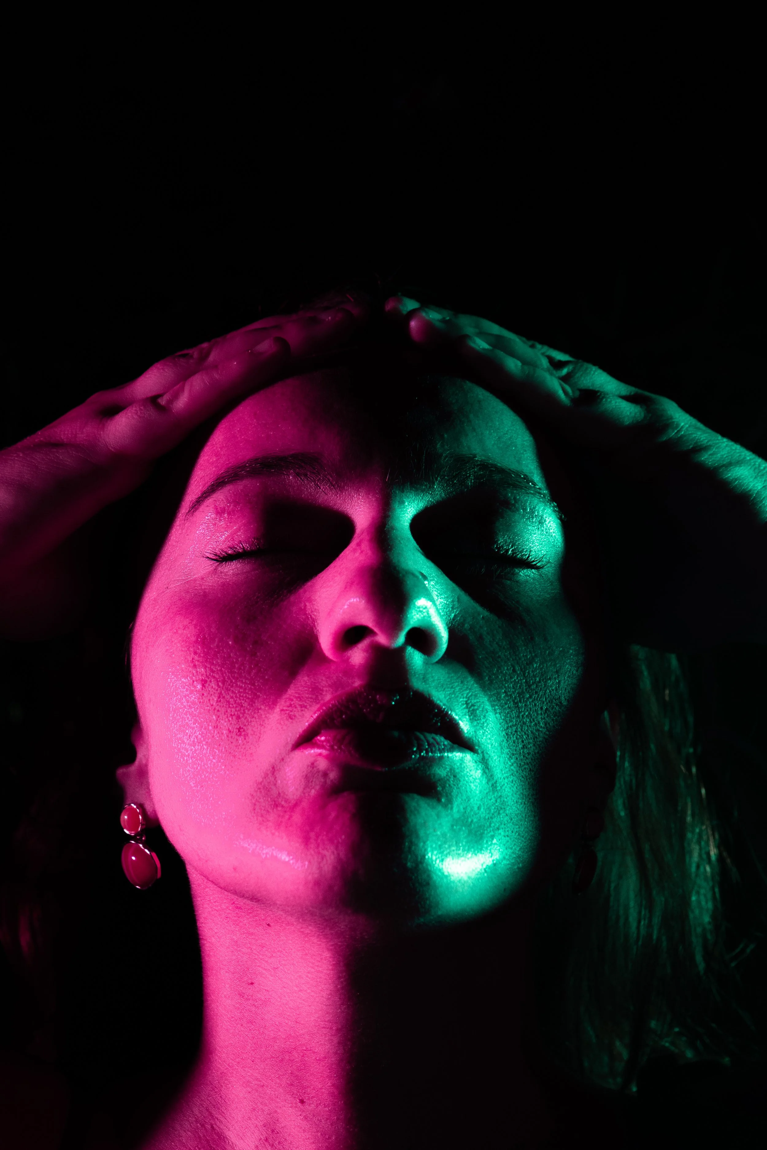 Portrait d'une femme avec visage éclairé en rose et vert, yeux fermés, main sur la tête, fond noir.
