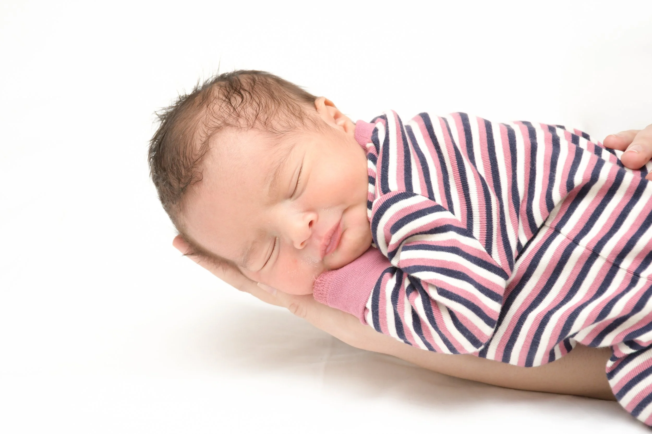 Un bébé endormi avec la tête posé dans la main d'une personne, portant un pyjama à rayures rose, gris et blanc, sur un fond blanc.