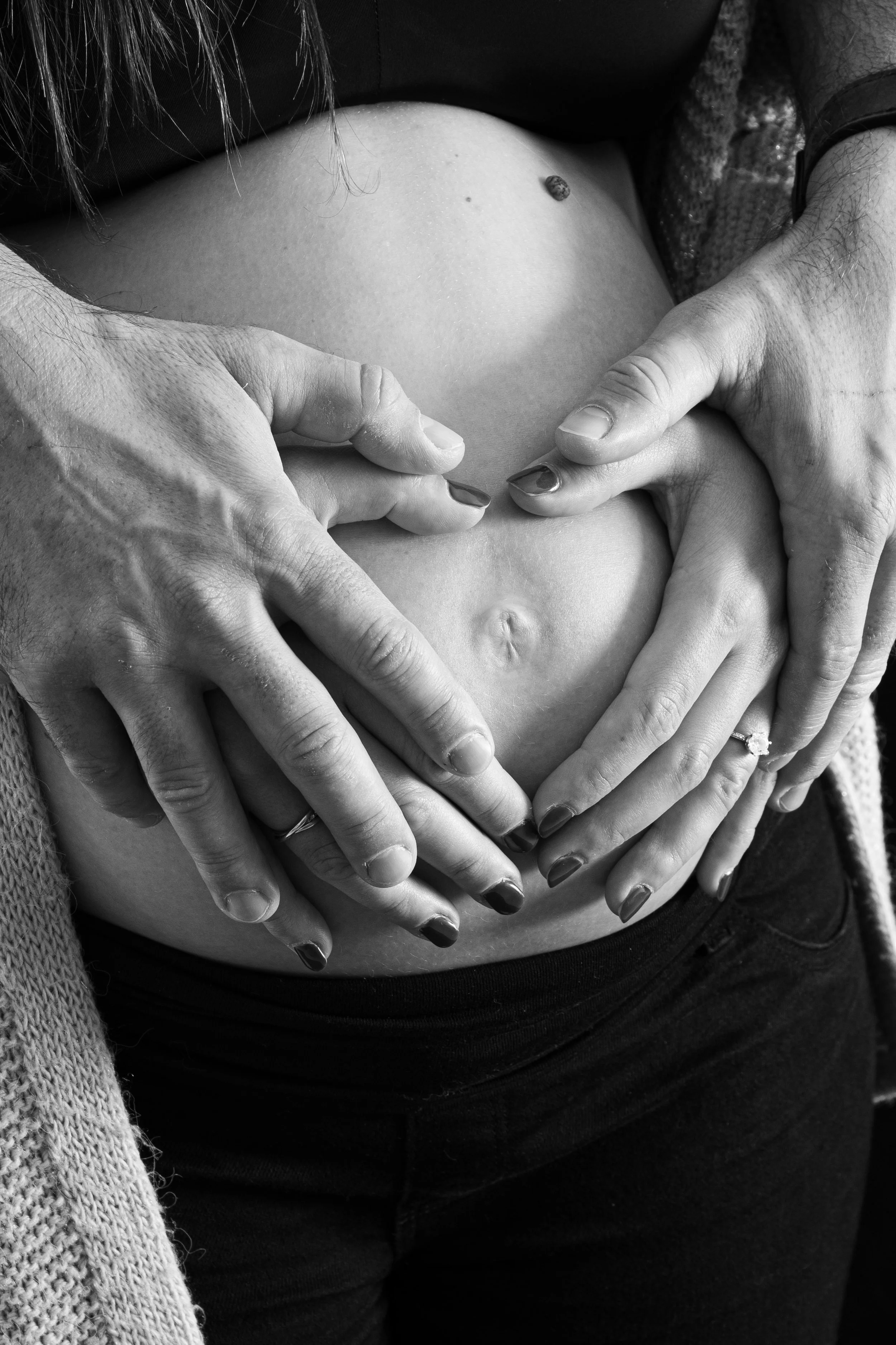 Une femme enceinte dont le ventre est entouré par plusieurs mains, formant un cœur, en noir et blanc.