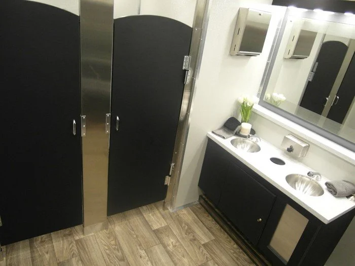Public bathroom with black and stainless steel stalls, a black and white sink area with two round sinks, a mirror, soap dispenser, black towels, a small vase with white flowers, and a wooden floor.
