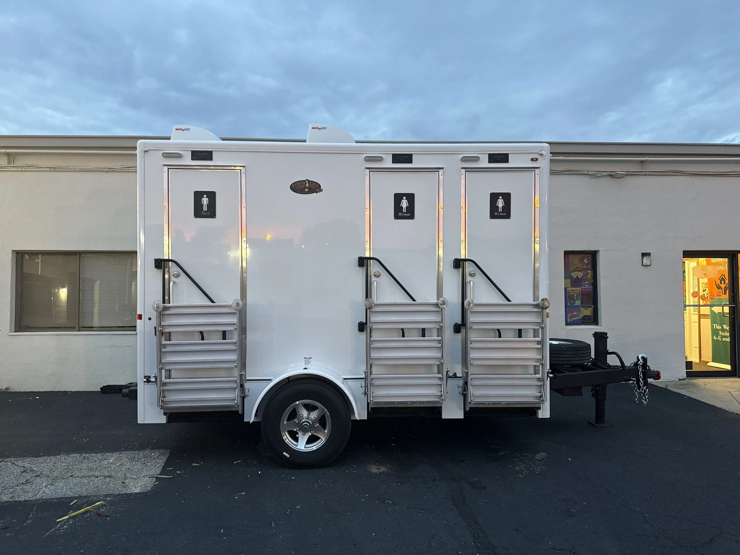 A white portable trailer with restroom signs for men and women parked outside a building with lit windows, at dusk, under a partly cloudy sky.