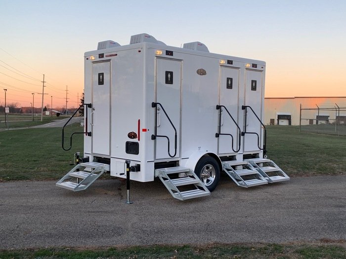 A white mobile restroom trailer with three separate entrances and fold-out steps, parked on a grassy area near a chain-link fence at sunset.