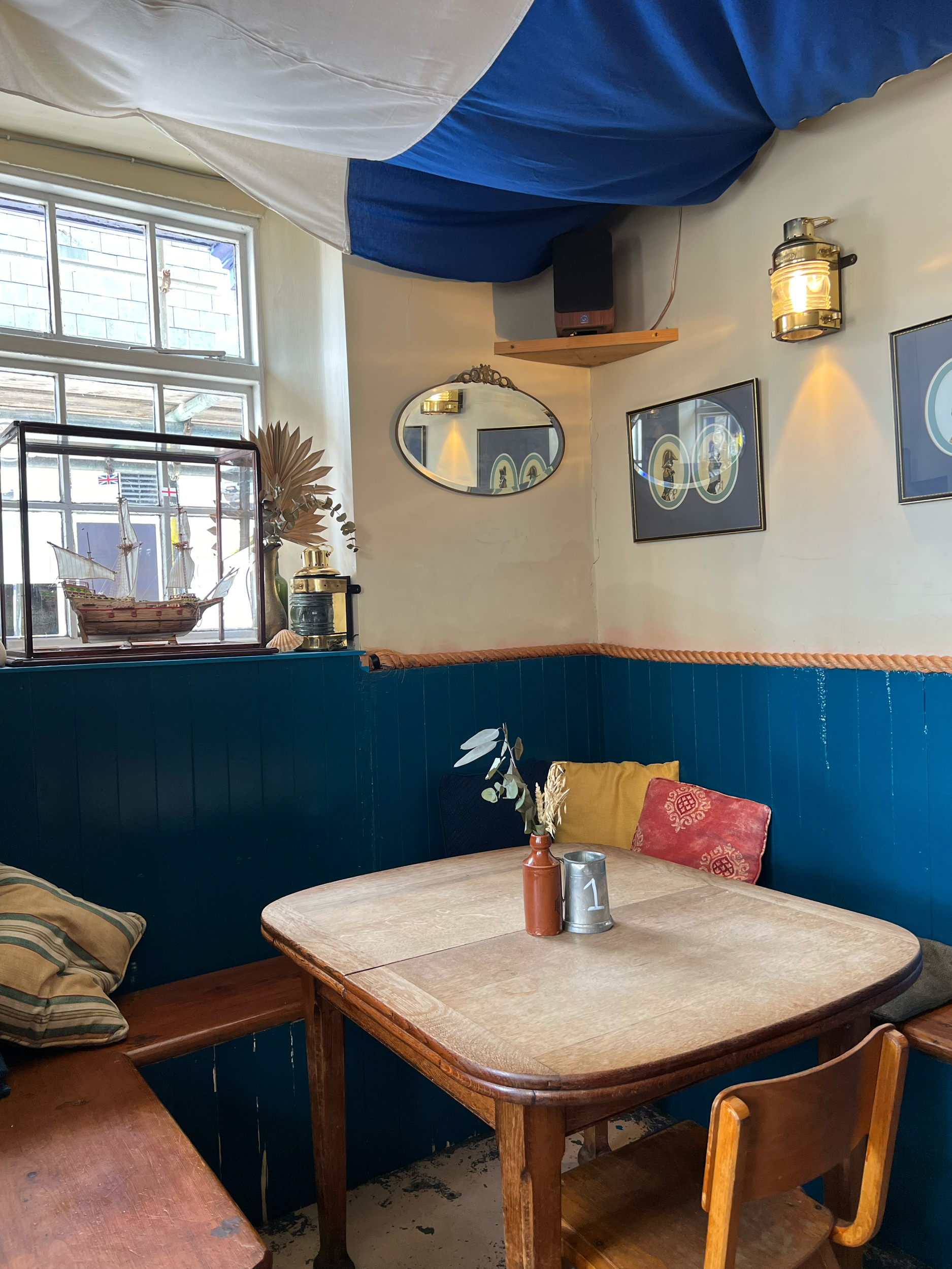 Cozy cafe interior with a wooden table covered with decorative items including vases and leaves, surrounded by mismatched wooden chairs, a window letting in natural light, and artwork on the walls.