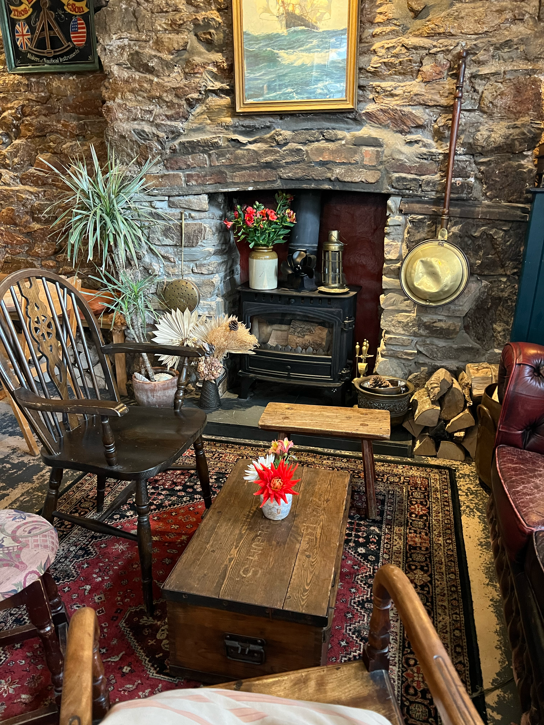 A cozy living room with a stone fireplace, wooden furniture, and decorative plants. The mantle holds ornaments and a framed painting, with a vase of pink flowers nearby. There is a small black stove and an area rug on the floor.