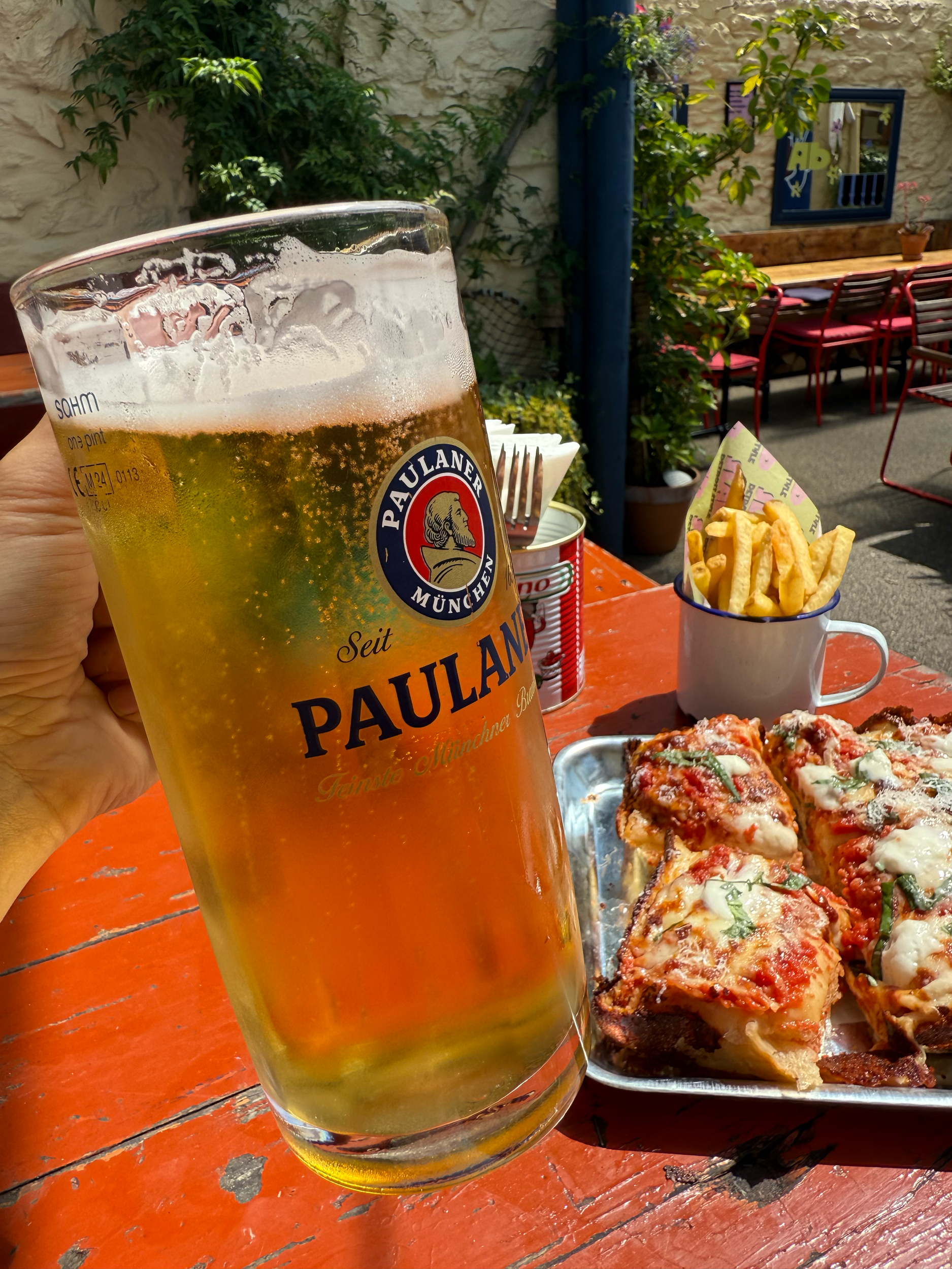 A glass of Paulaner beer, a tray of lasagna, a cup of french fries, and a can of soda on an outdoor wooden table with a person holding the beer.