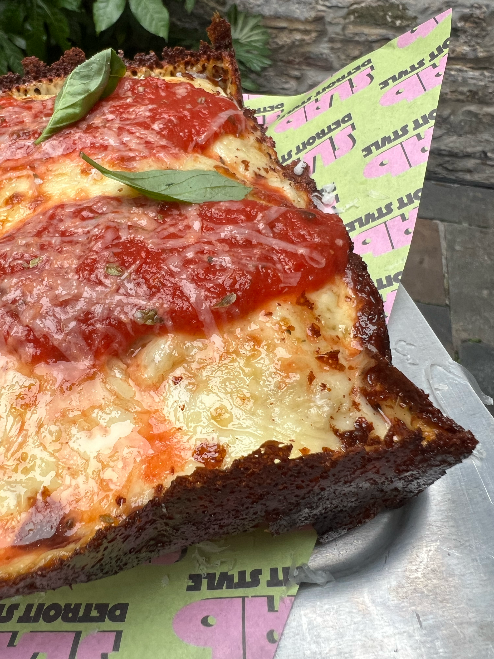 A slice of pizza with cheese, tomato sauce, basil leaves, and a thick, dark crust, served on a green and pink paper liner.
