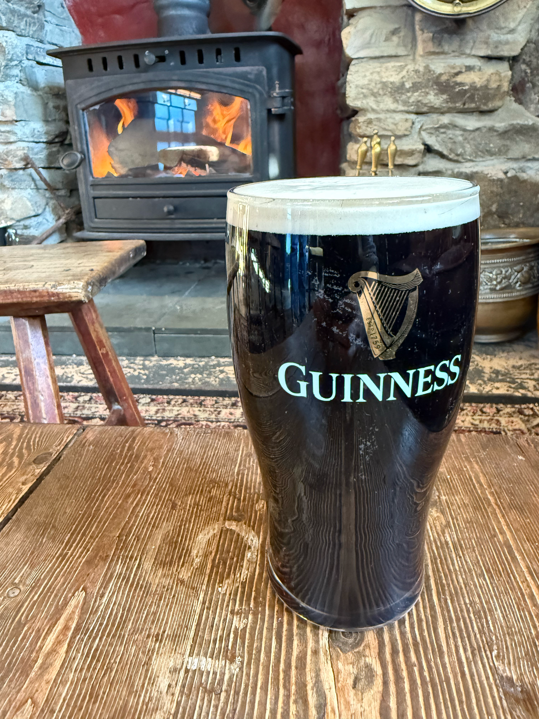 A large Guinness beer glass with a Guinness logo, filled with dark stout, placed on a wooden table in front of a stone fireplace and traditional wood stove.