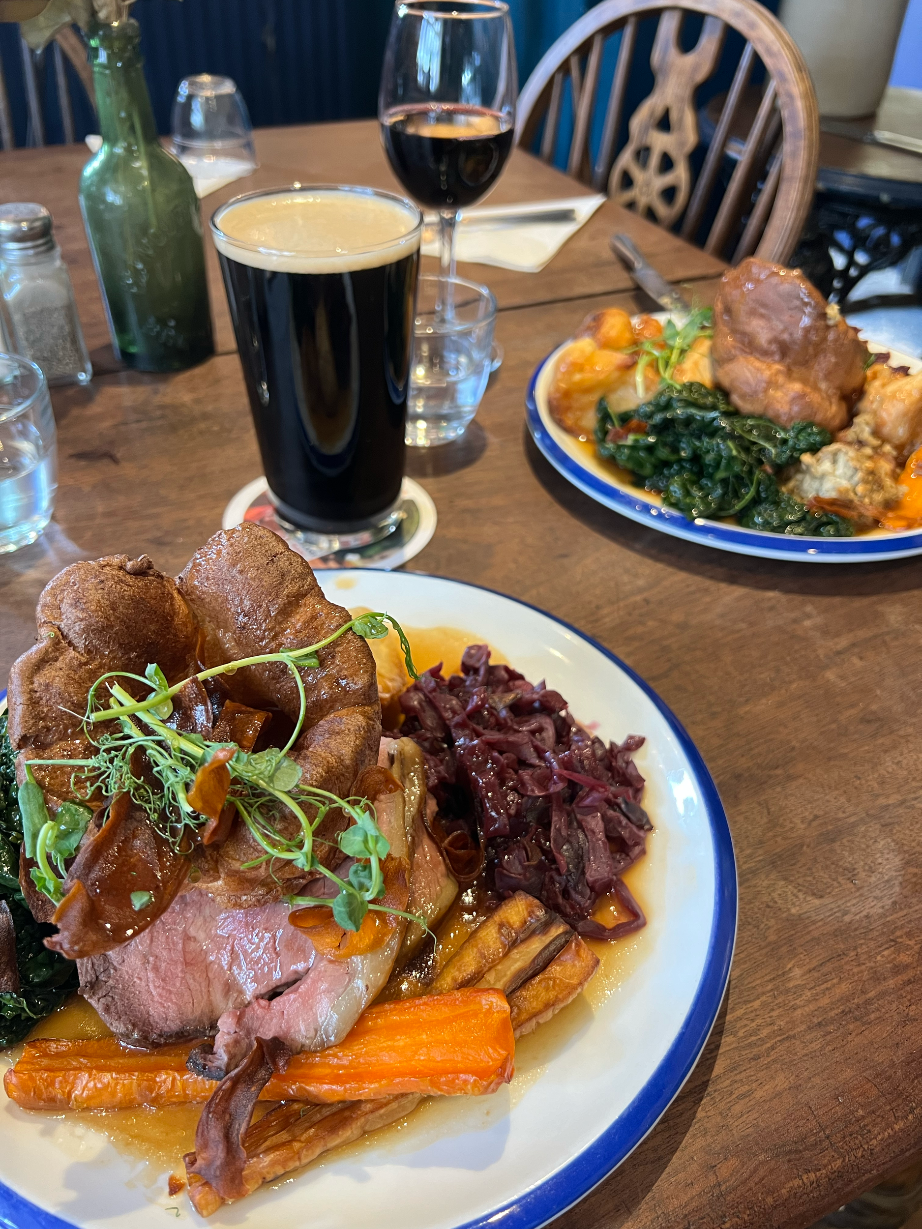 A plate of carved beef roast with vegetables like carrots and a side of red cabbage, garnished with microgreens, served with two glasses of dark soda and a pint of dark beer. In the background, there is another plate with food and several glass bottl