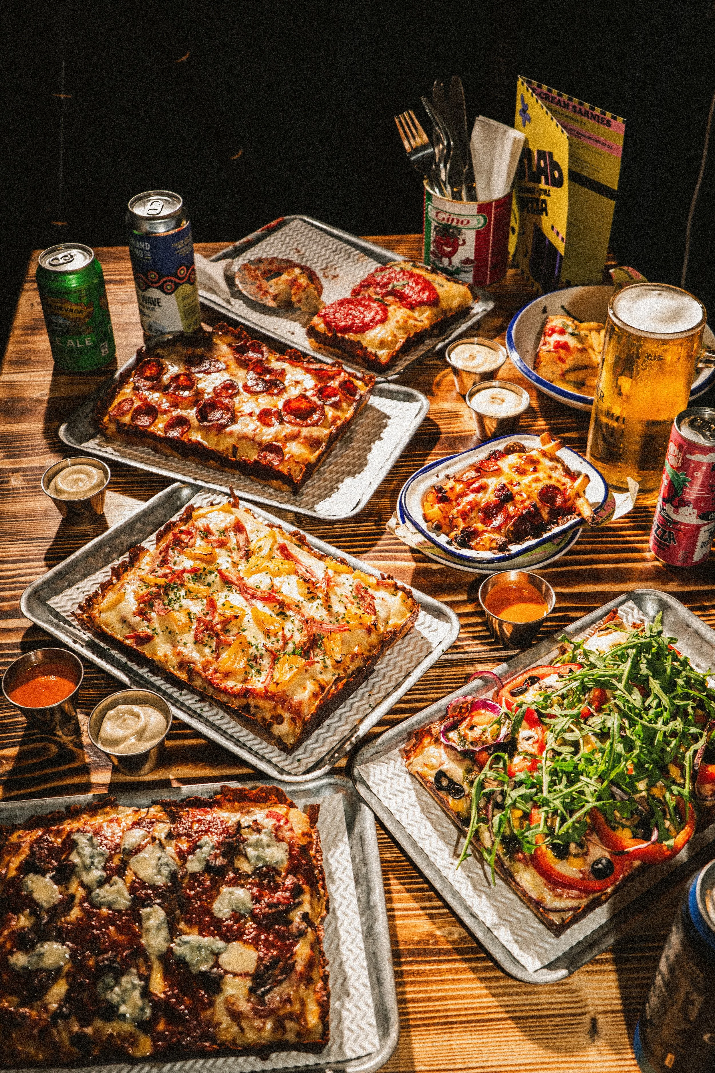 A variety of pizzas on metal trays, a salad with arugula, and beverages including beer cans and shots on a wooden table.