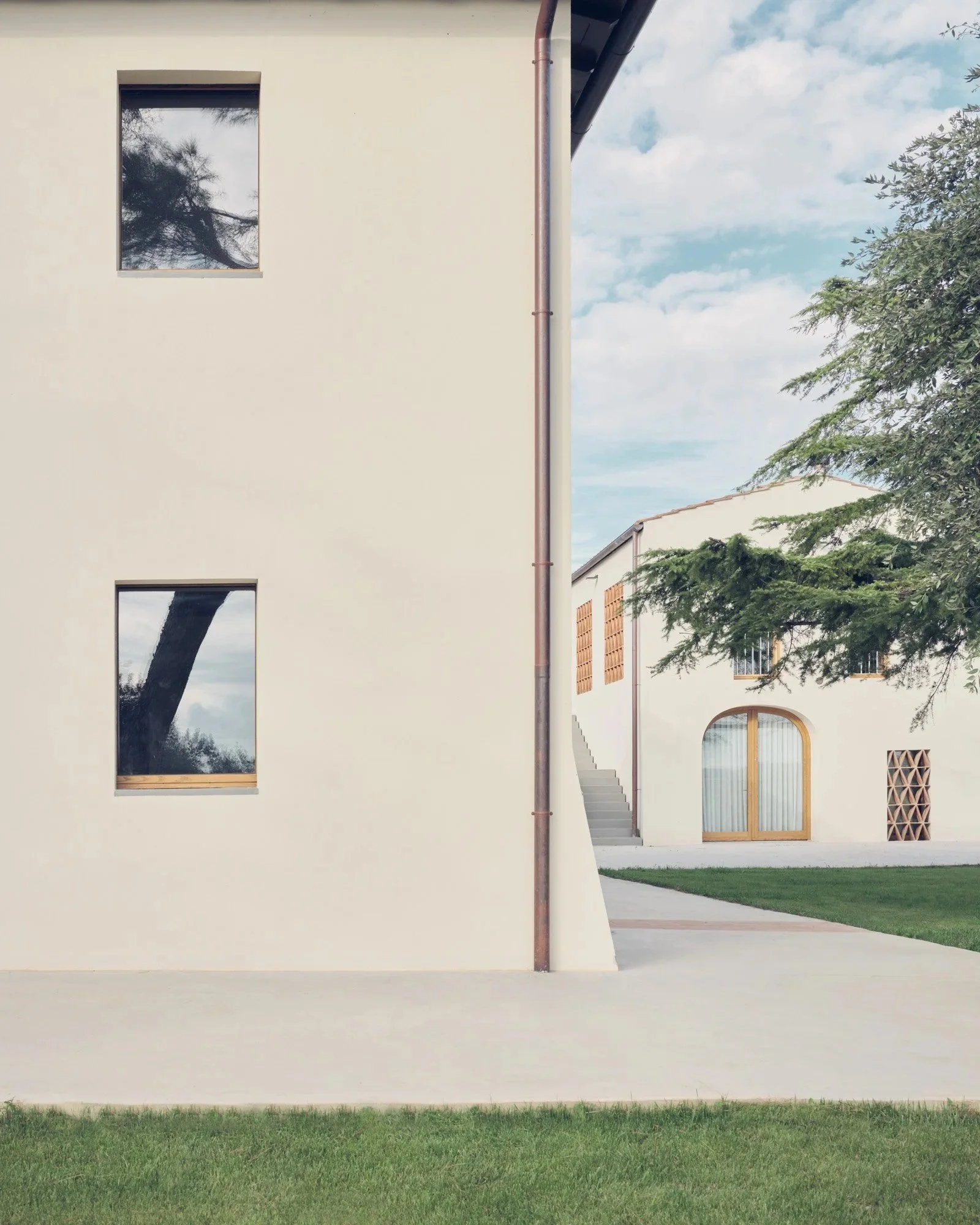 Parte di un edificio moderno con pareti bianche, finestre moderne e un pavimento di cemento, con alcune aree di prato verde e un albero a destra.