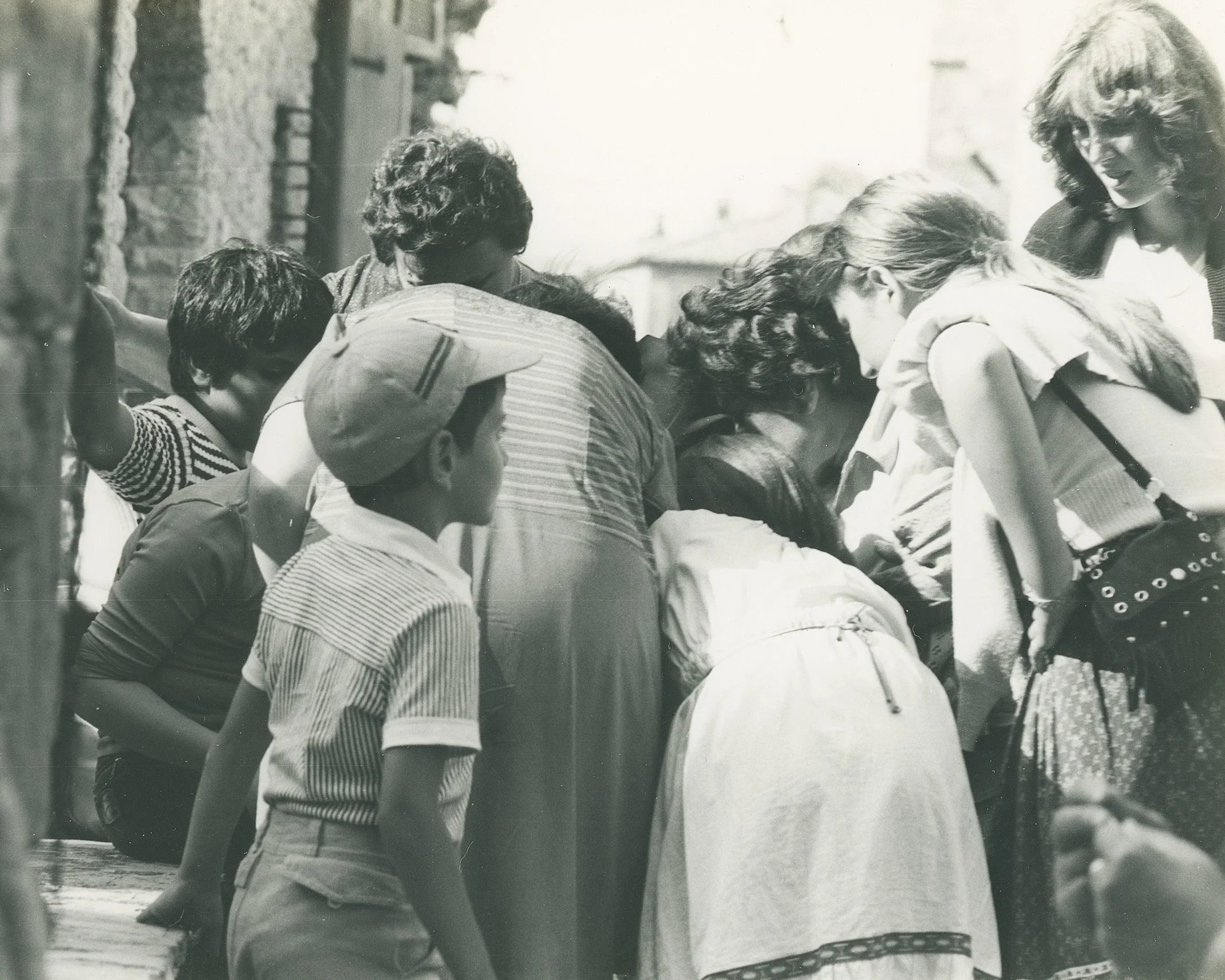 Un gruppo di persone, tra cui adulti e bambini, si raccoglie molto vicino e sembra osservare o partecipare a qualcosa di interessante, forse un evento o un oggetto sotto attenzione. L'immagine è in bianco e nero.