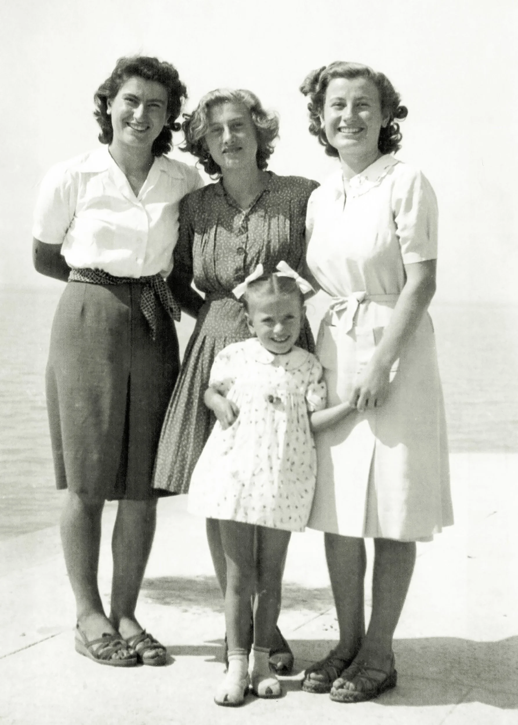 Foto in bianco e nero di quattro donne adulte e una bambina sorridenti, in spiaggia, con il mare sullo sfondo.