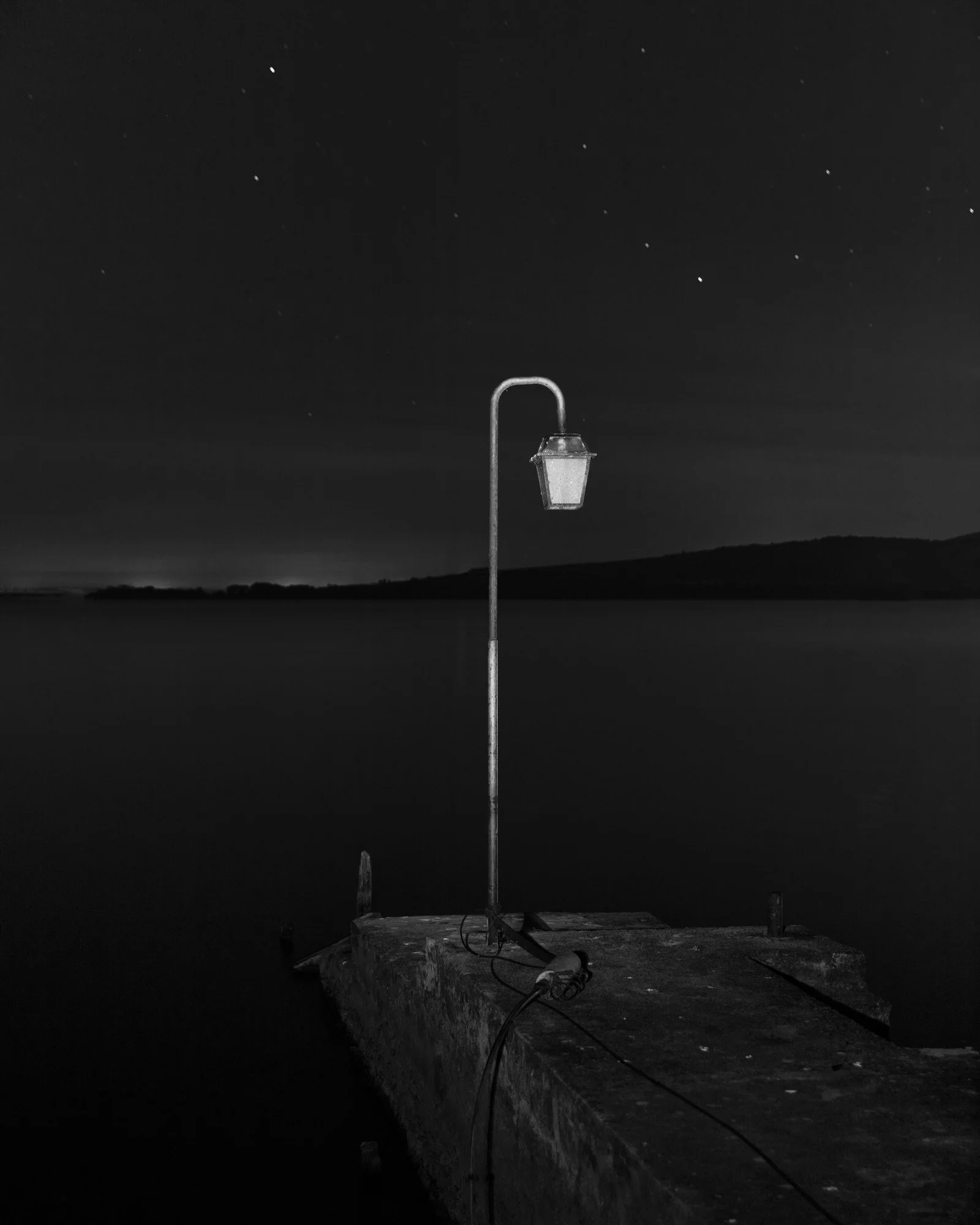 Lampione su un molo di pietra di notte con cielo stellato e silhouette di colline sullo sfondo.