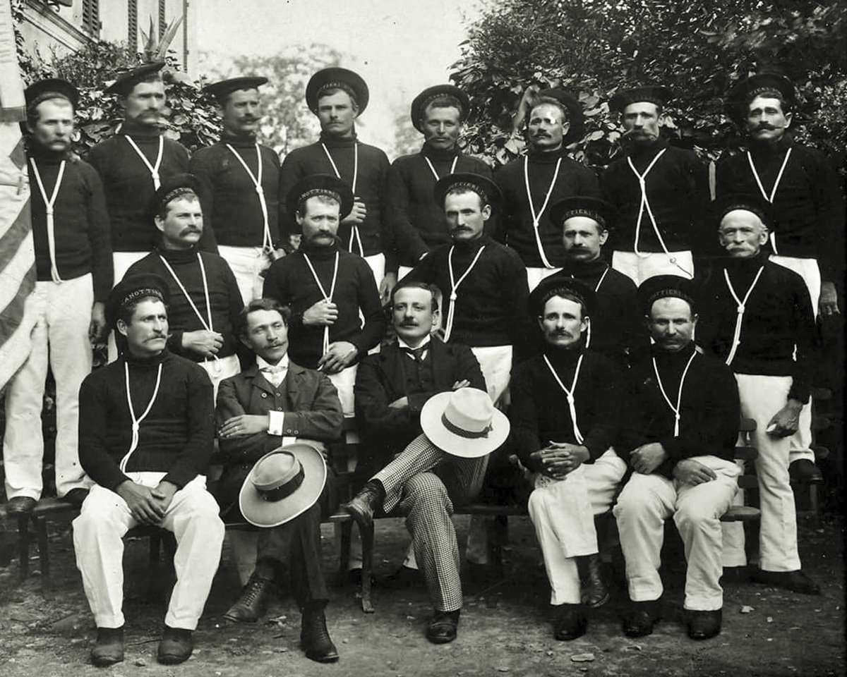 Gruppo di uomini in uniforme con cappelli, alcuni seduti e altri in piedi, in un ambiente esterno con vegetazione sullo sfondo, foto in bianco e nero.