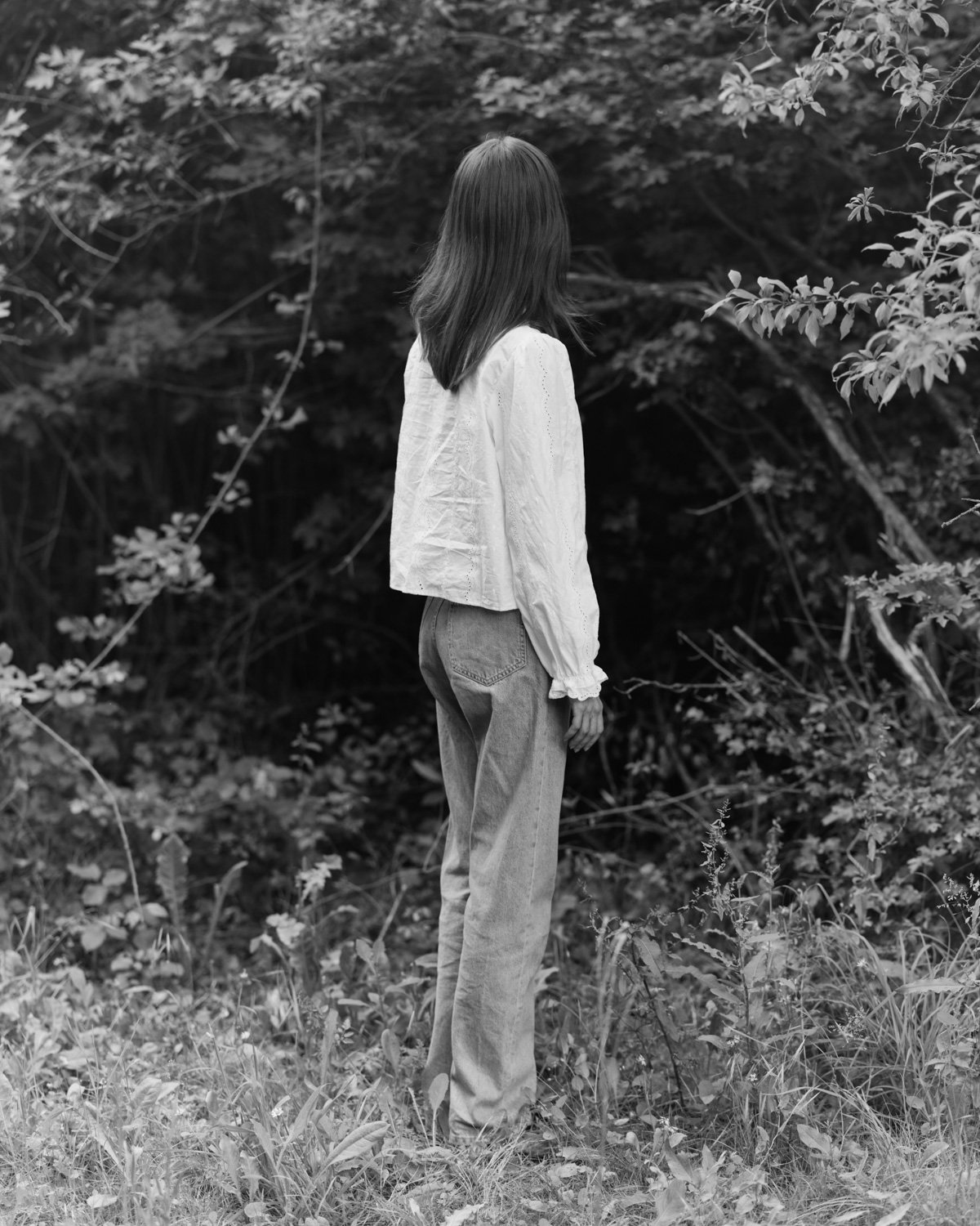 Una donna vista di spalle indossa una camicetta bianca e jeans, in mezzo alla natura con foglie e rami attorno, in bianco e nero.