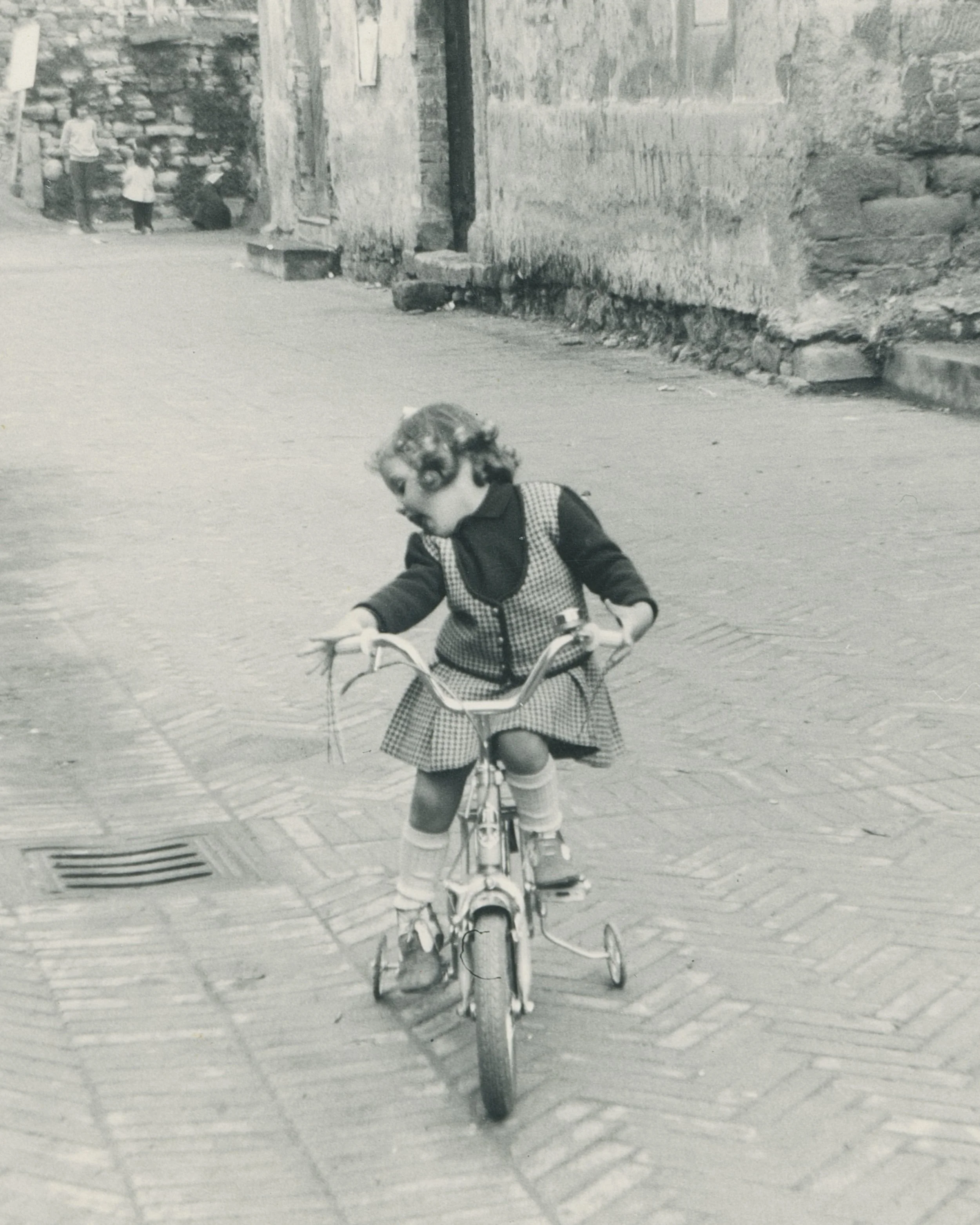Una bambina con un vestito a quadri pedala su una bicicletta con le ruote di supporto in un vicolo di pietra in bianco e nero.