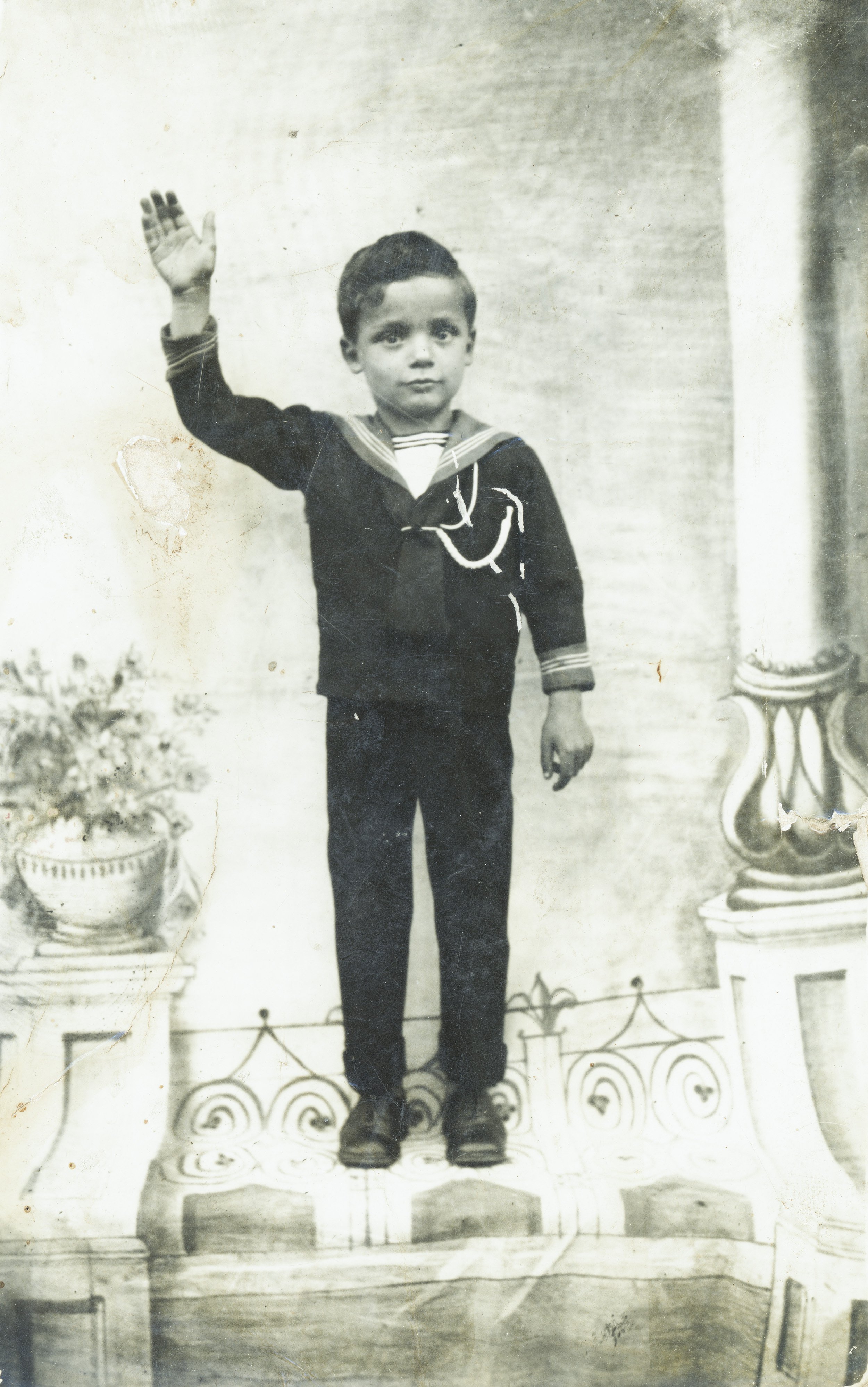 Bambino vestito in uniforme, in piedi su un grande disegno dipinto, che saluta con la mano destra, in un'ambientazione di scena.