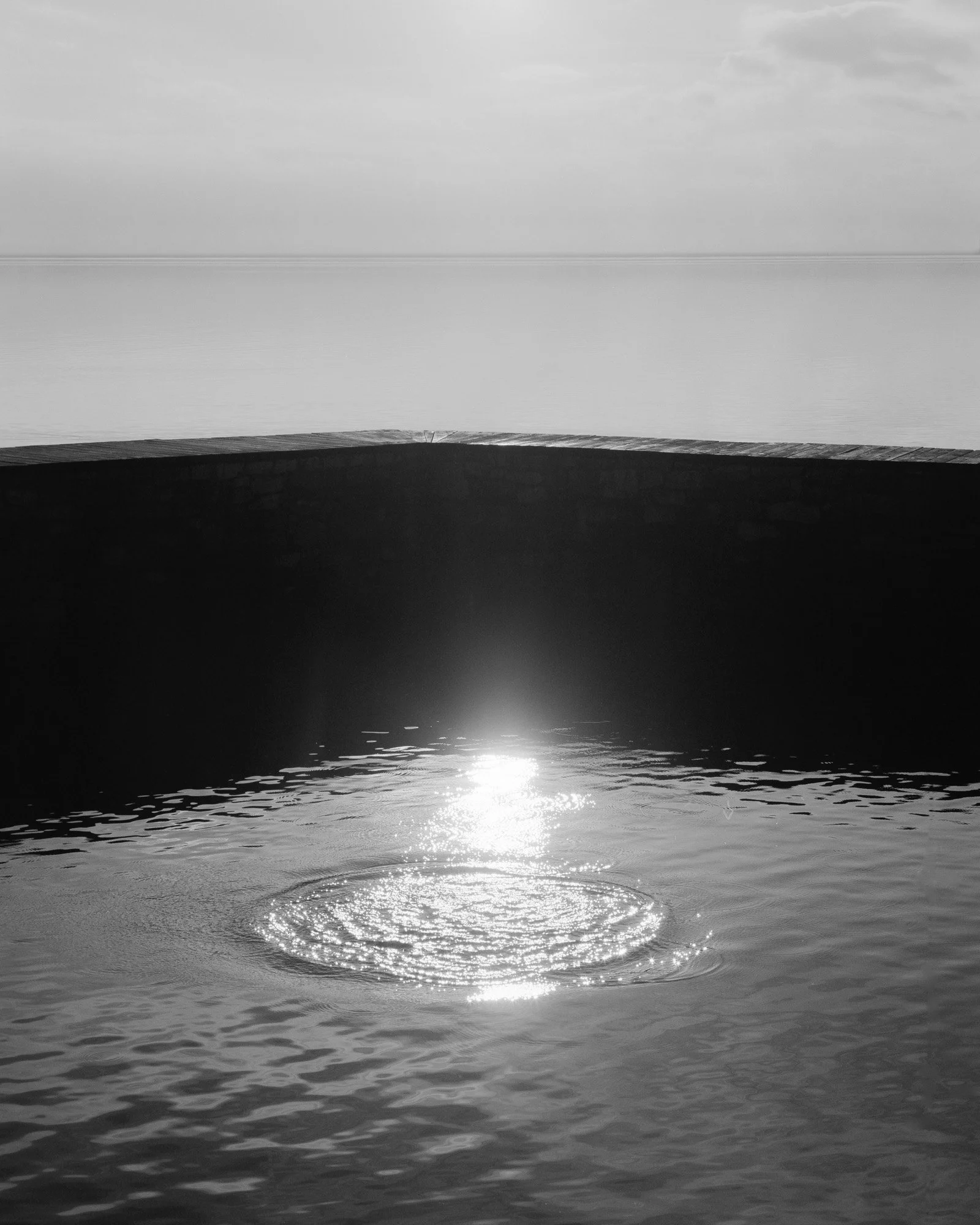 Foto in bianco e nero di un paesaggio marino con acqua calma, un molo di legno, e il riflesso del sole sulla superficie dell'acqua.