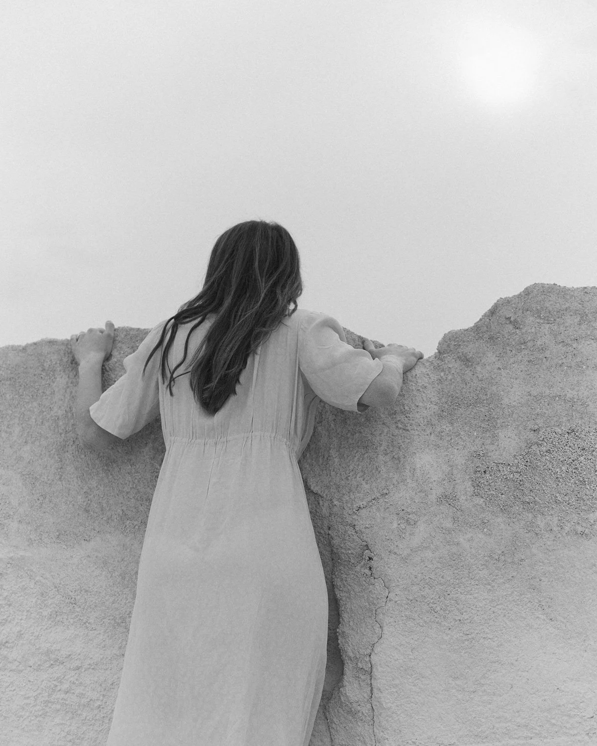 Donna con capelli lunghi che si appoggia a un muro di pietra, vista di schiena in bianco e nero.
