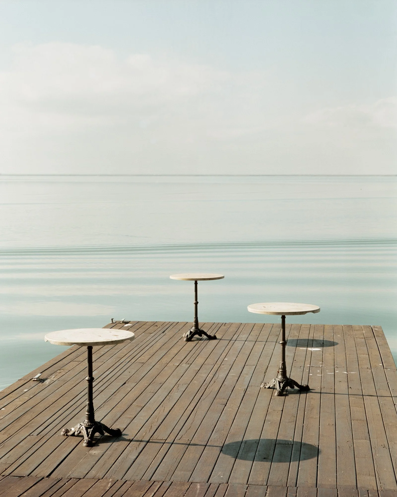 Tre tavolini rotondi su un pontile di legno che si affaccia su un mare calmo, cielo con poche nuvole.