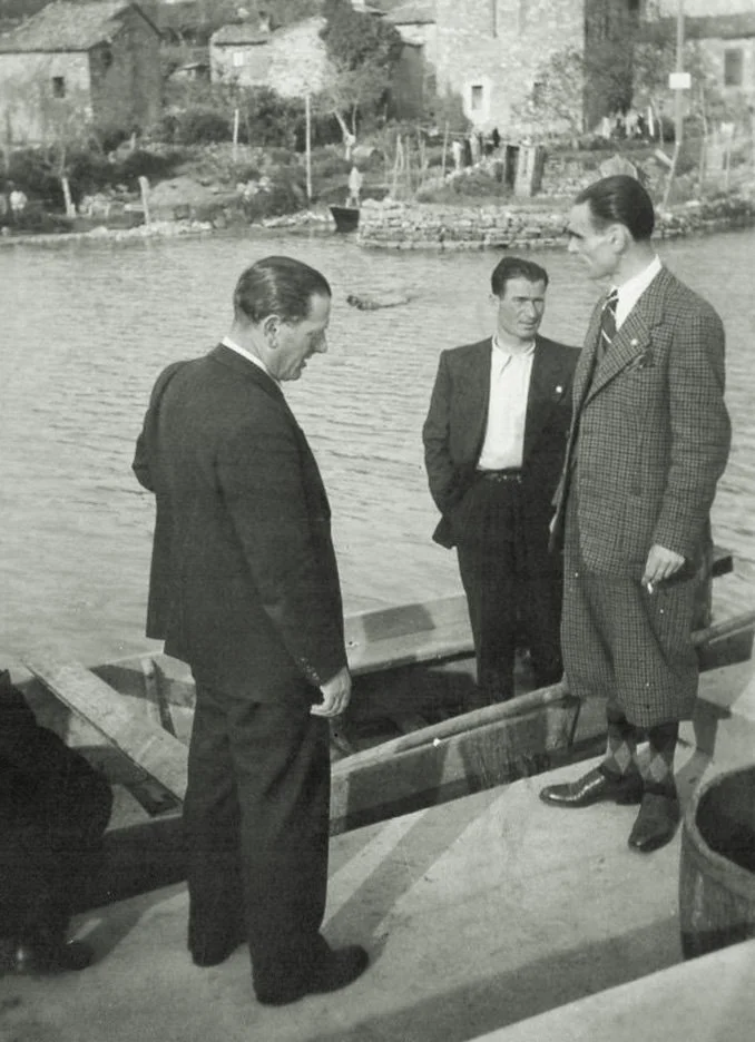 Tre uomini in abiti formali sono vicino a un fiume o un lago, con sfondo di case e vegetazione. Sono impegnati in una conversazione, con uno di loro che guarda verso l'acqua.