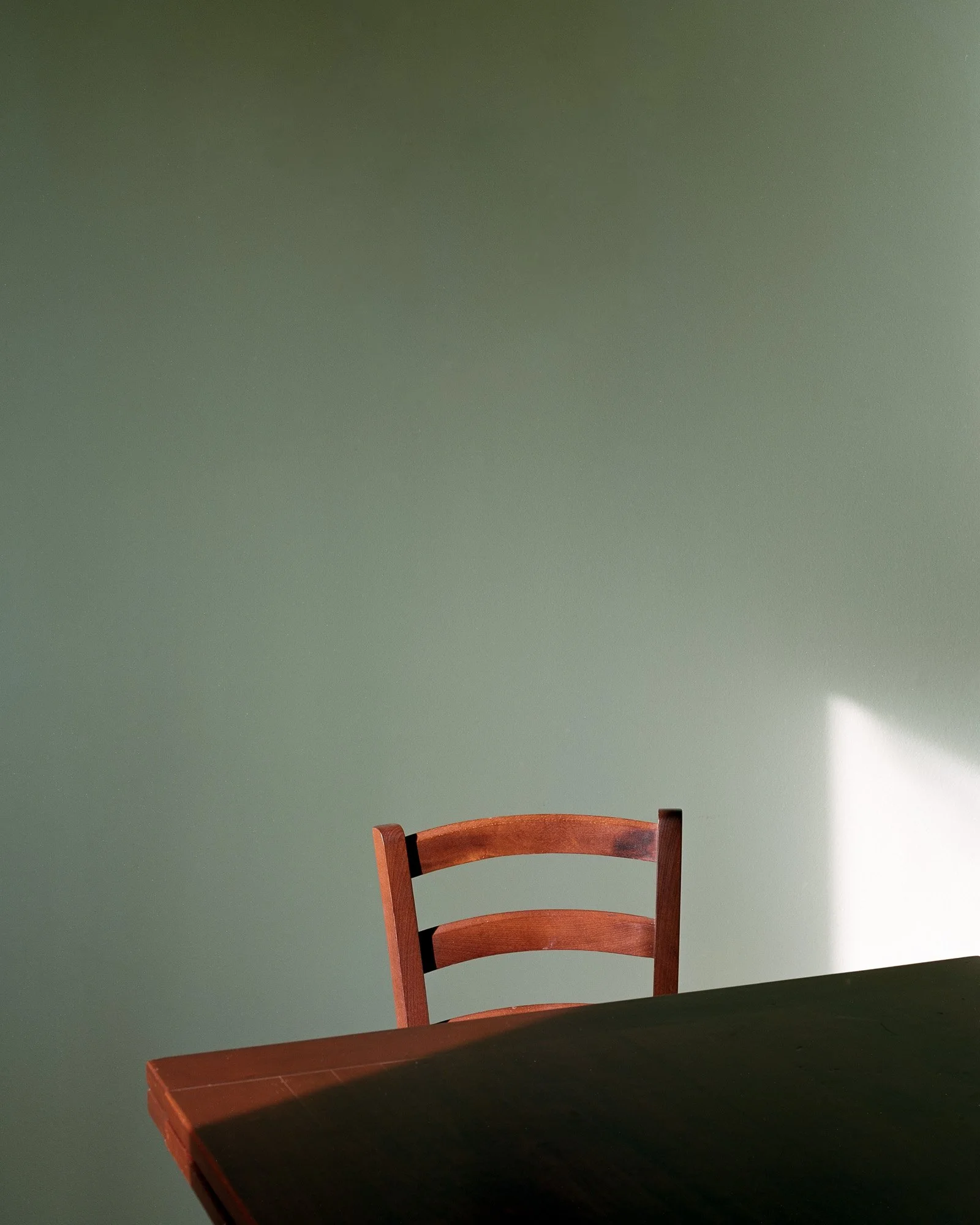 Una sedia di legno davanti a un tavolo scuro, con sfondo verde e luce che entra da destra.