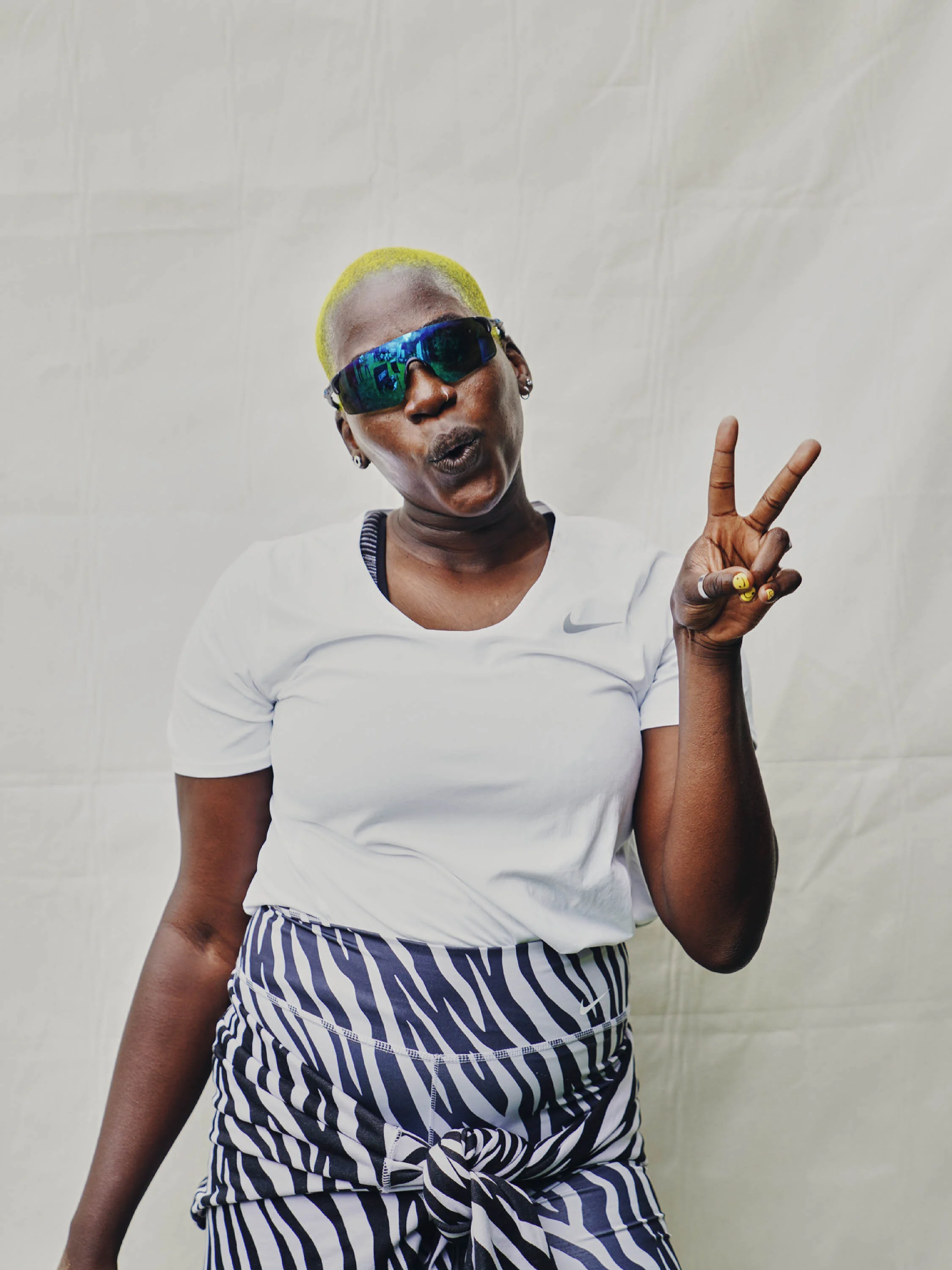 A woman with short yellow hair wearing sunglasses, a white Nike t-shirt, and a black-and-white striped skirt making a peace sign.
