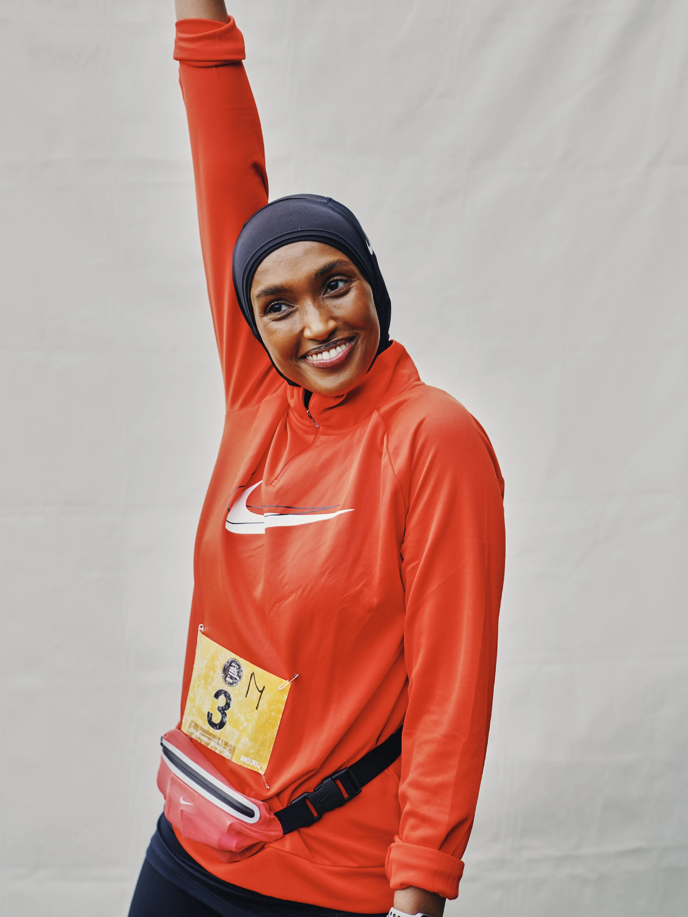 A woman wearing a black hijab, orange Nike sports jacket, and black pants, smiling, with a yellow race bib numbered 3 and MY pinned to her jacket.