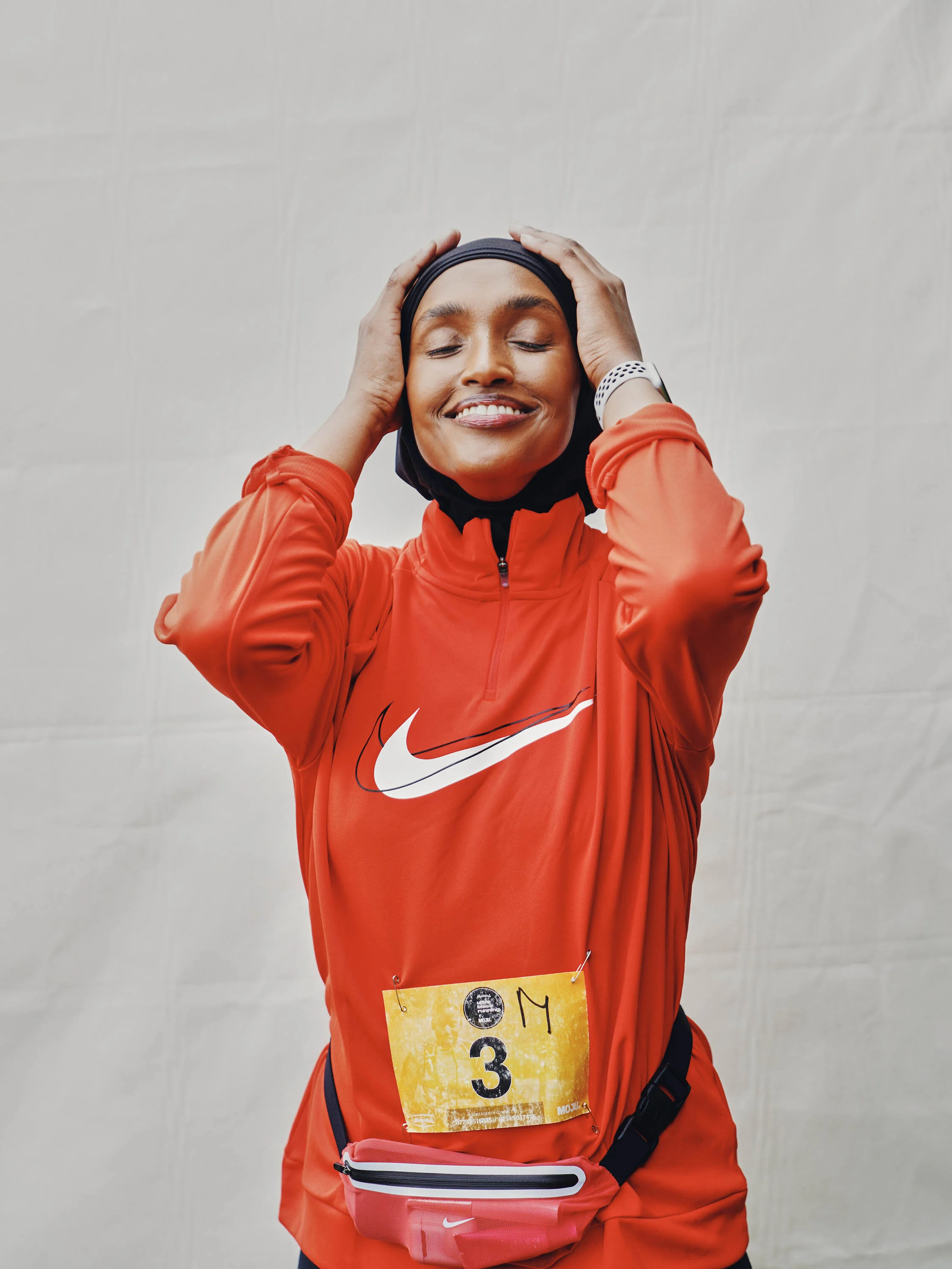 A woman wearing a black headscarf and red Nike athletic jacket, smiling with her eyes closed, holding her head with both hands, standing against a plain white wall