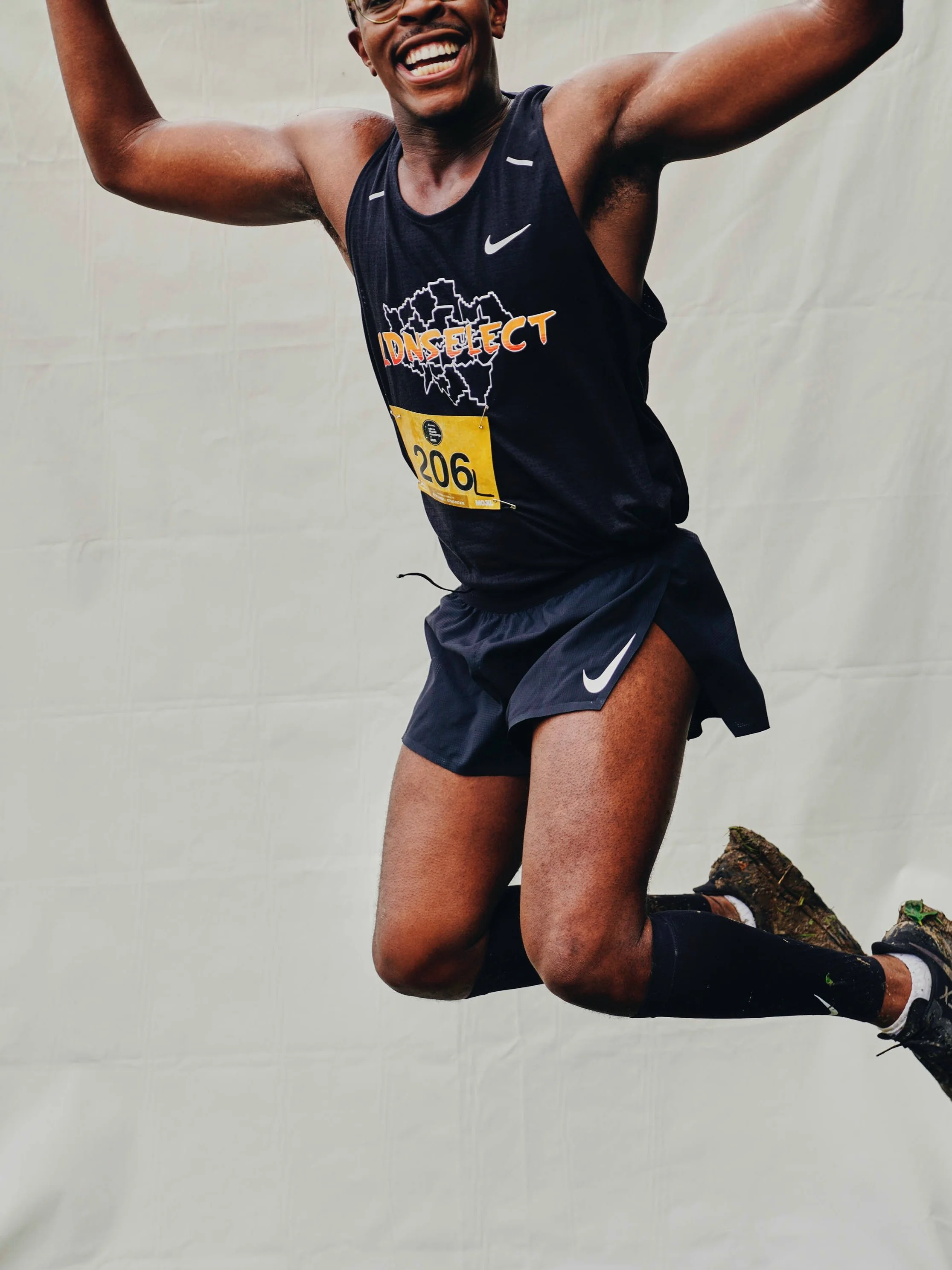 A male runner mid-air during a race, smiling and wearing a black sleeveless shirt with a graphic and the word 'INJECT,' black shorts, and running shoes.