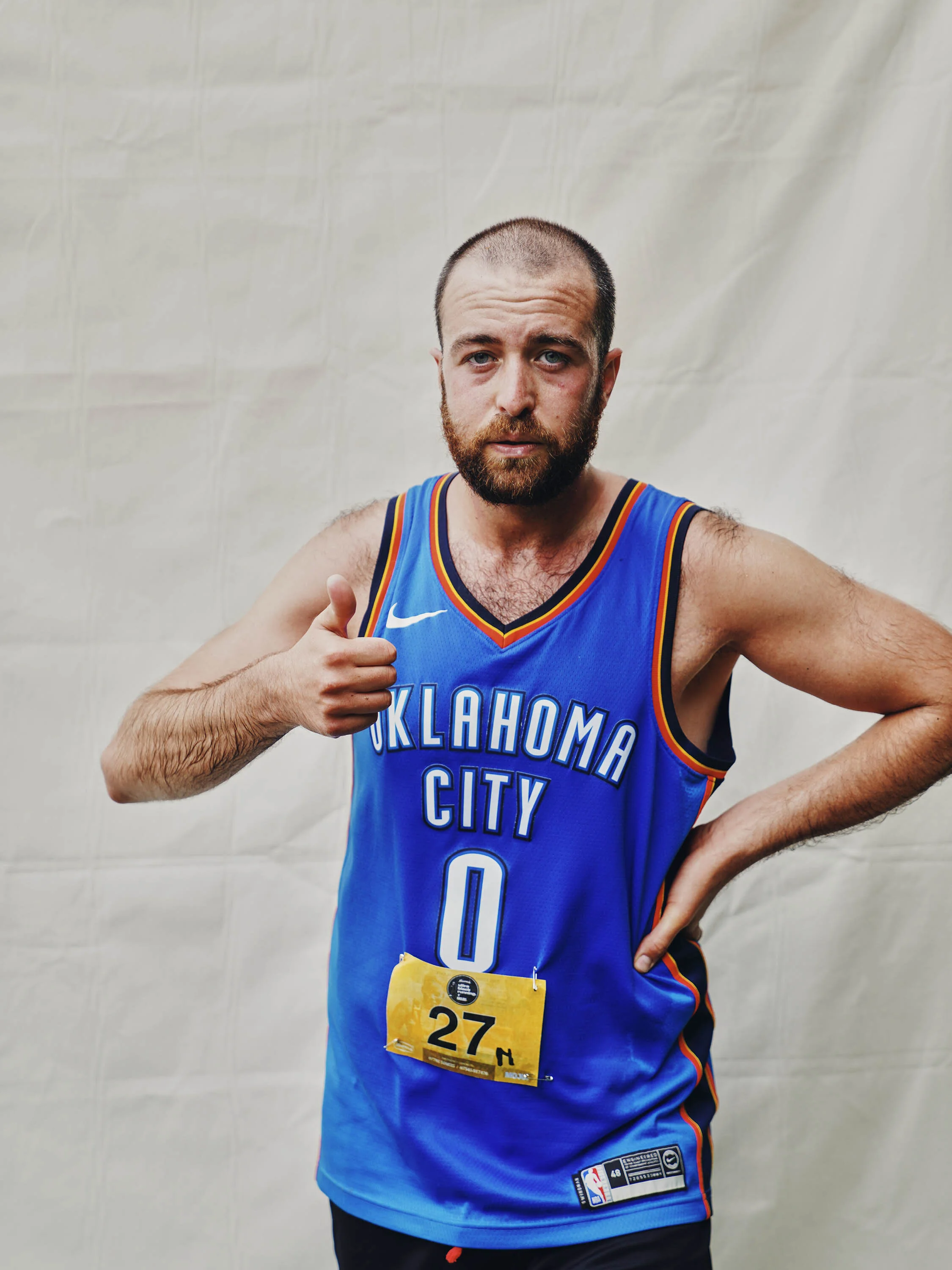A man with a beard wearing a blue Oklahoma City basketball jersey, giving a thumbs-up, against a plain background.