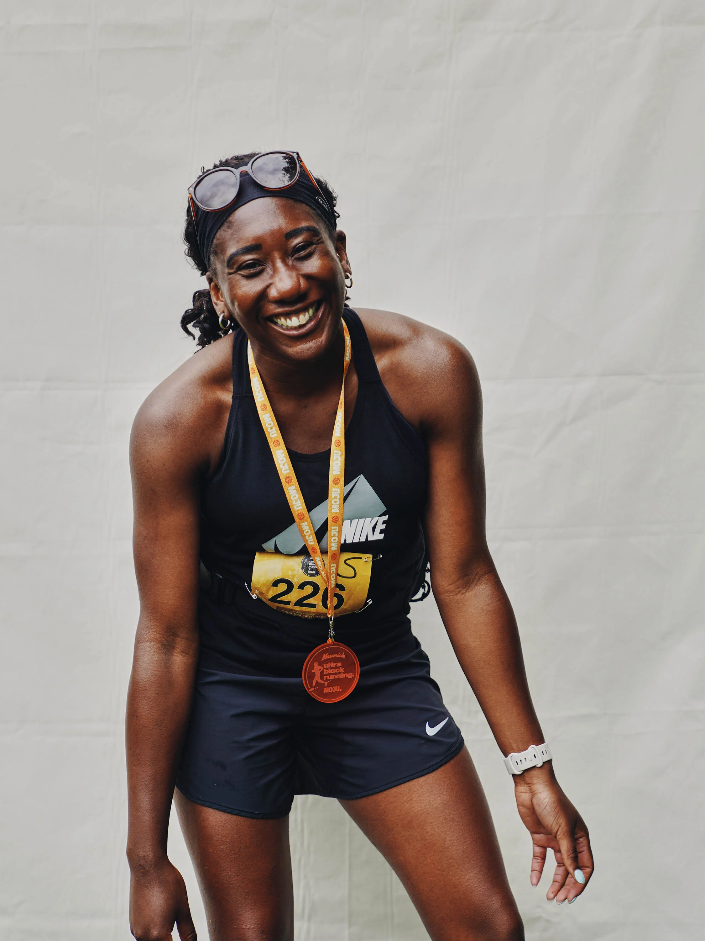 Female marathon runner smiling, wearing a black Nike tank top, black shorts, a white watch, a medal around her neck, sunglasses on her head, and a race bib number 226, standing against a plain background.
