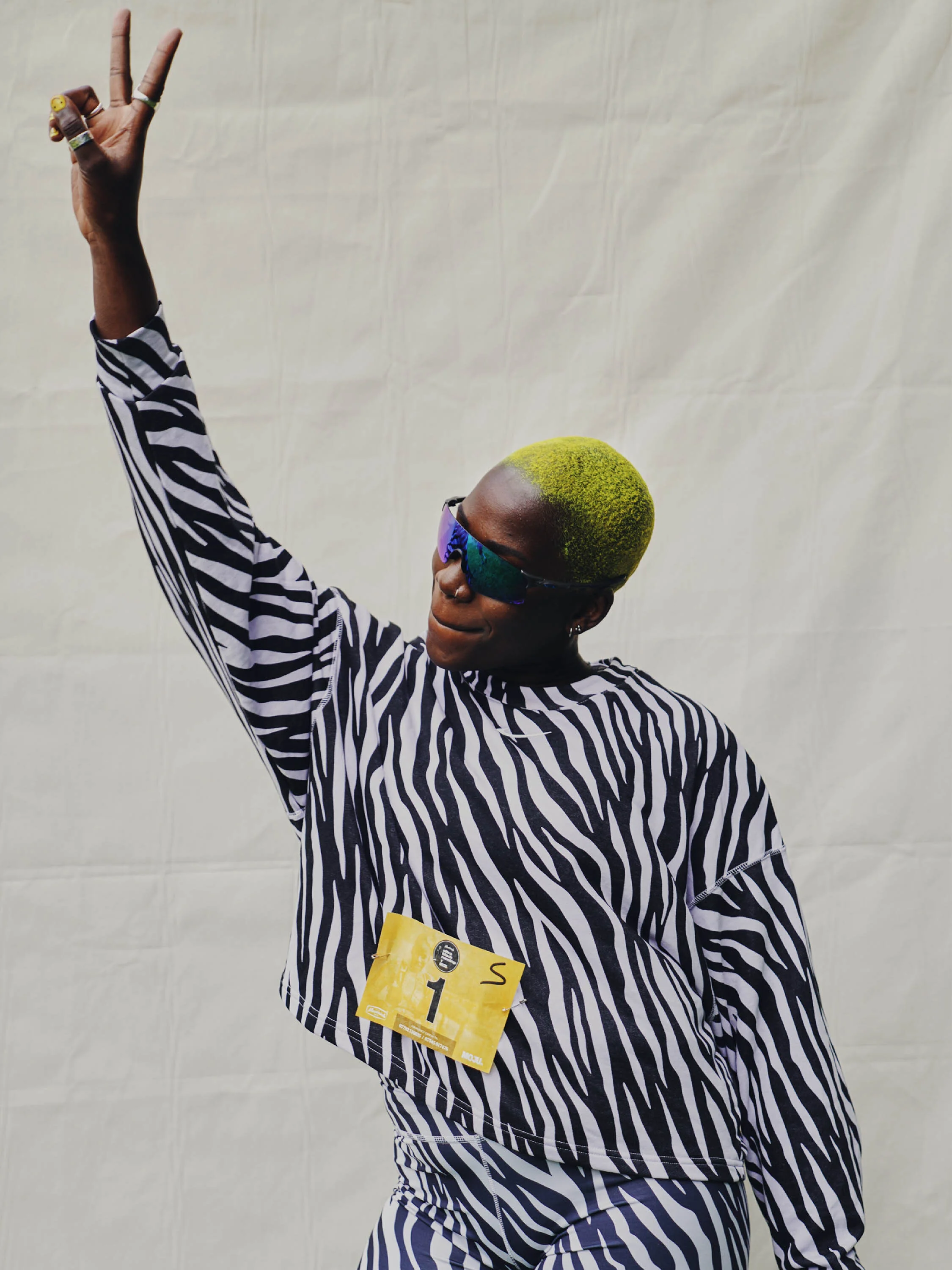 Person with lime green hair, wearing zebra print clothing and sunglasses, making a peace sign pose.