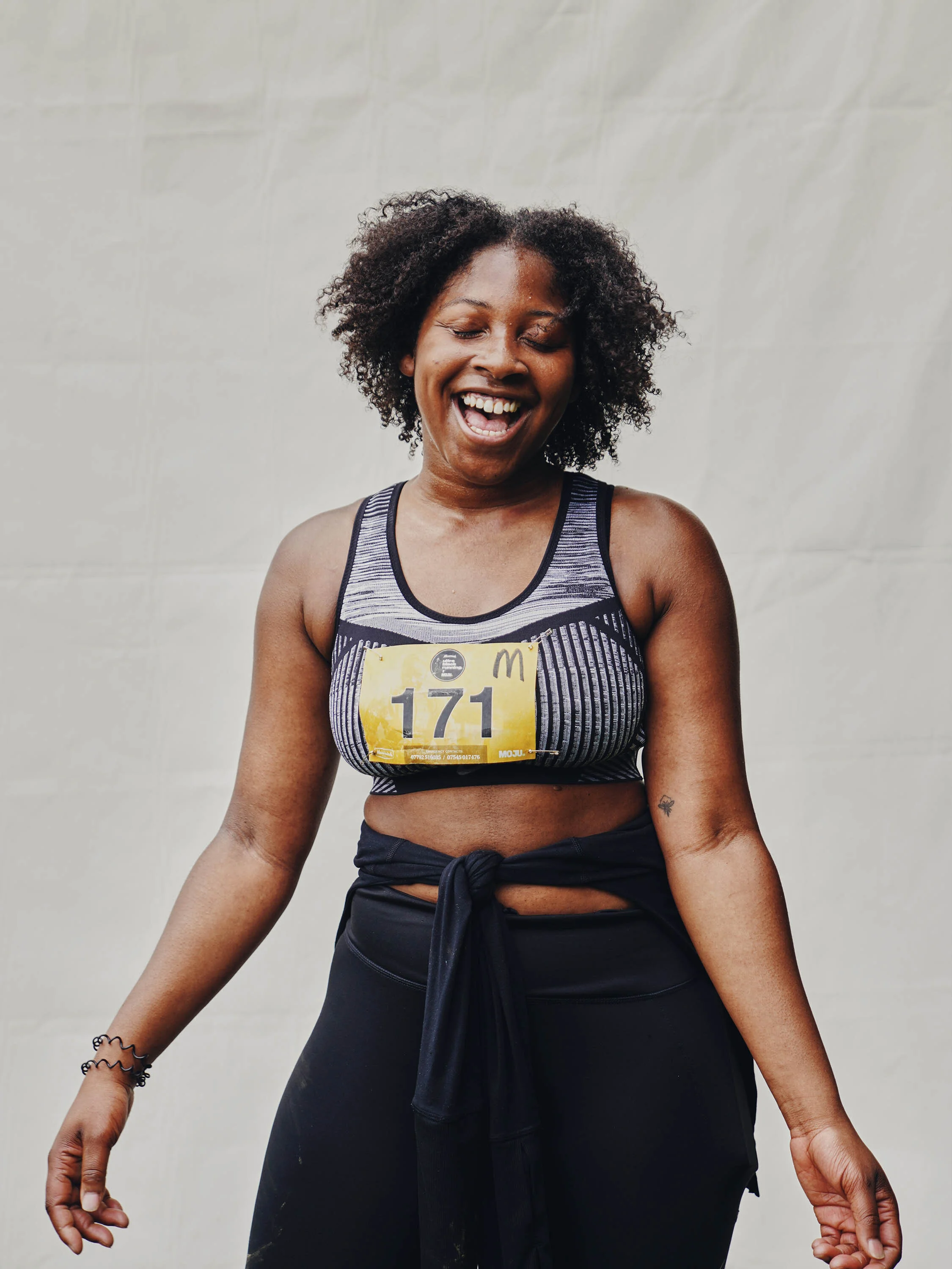 A woman with curly hair, wearing a striped sports bra and black leggings, smiling and looking down, with a yellow race bib number 171.