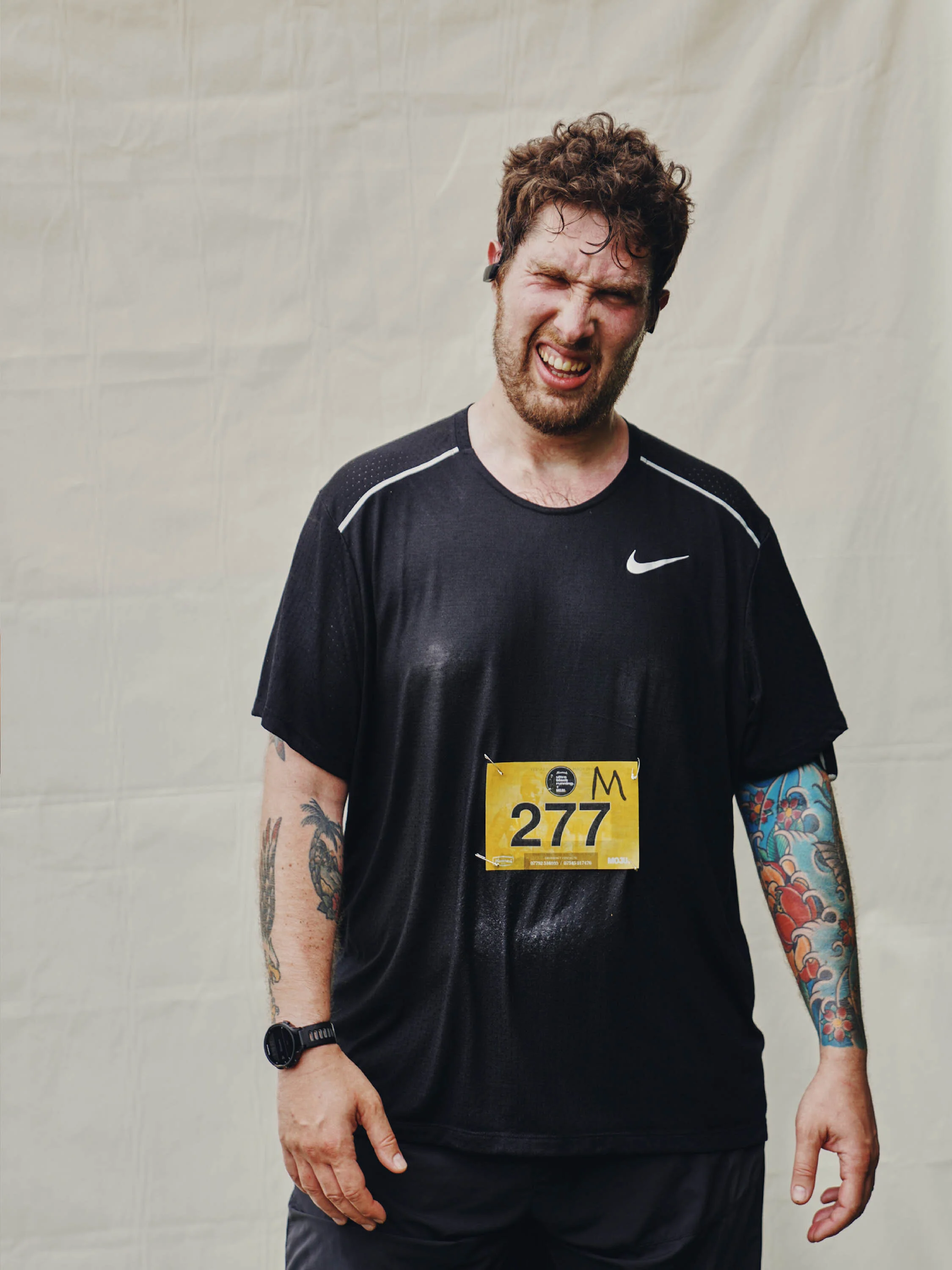 A man with messy brown hair and tattoos on both arms, wearing a black Nike running shirt with a race bib number 277, grimacing or making an uncomfortable facial expression, standing against a plain background.