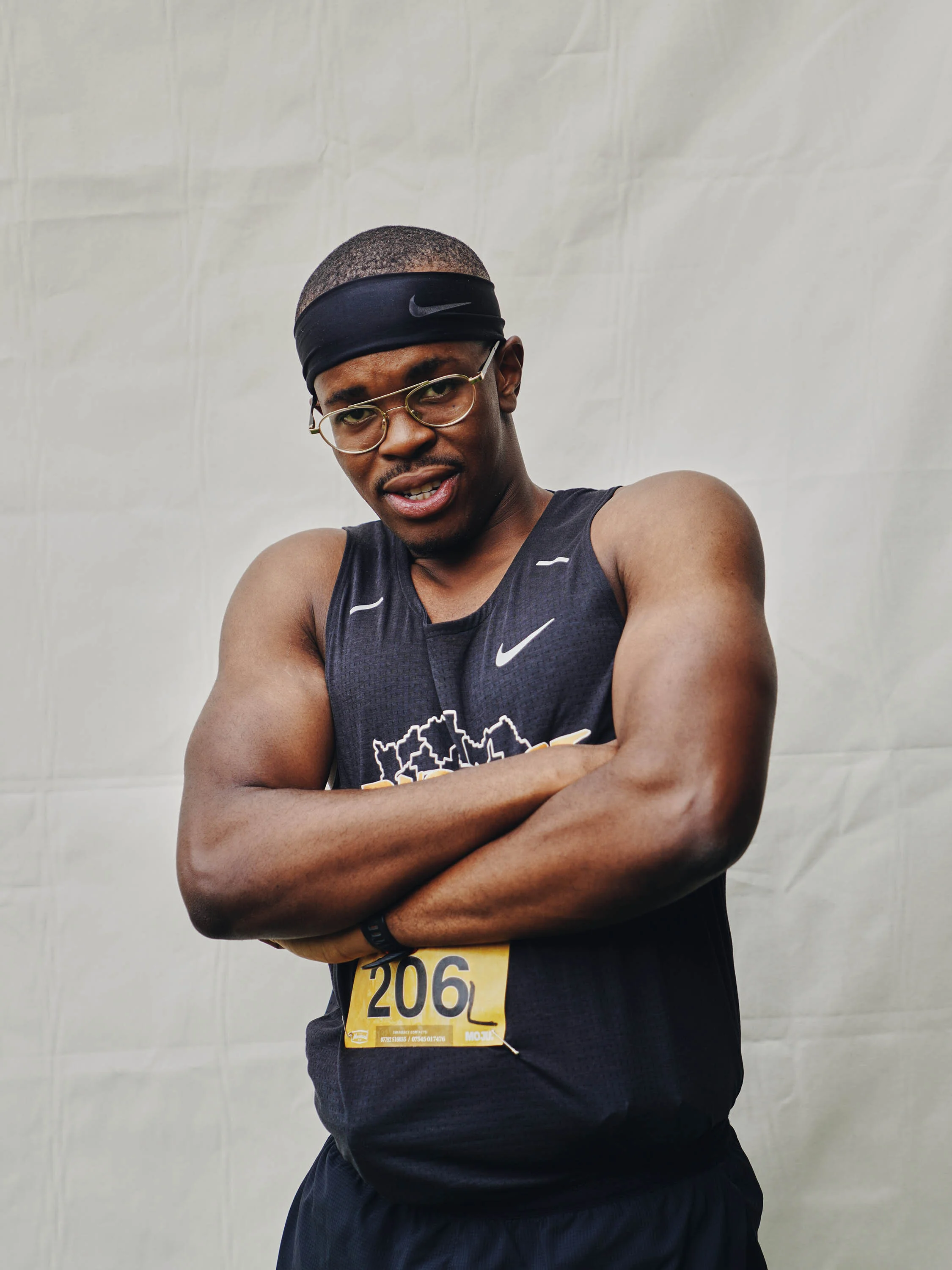 A young male athlete in black athletic wear and glasses, wearing a black headband, stands with arms crossed against a plain background, during a race or marathon.
