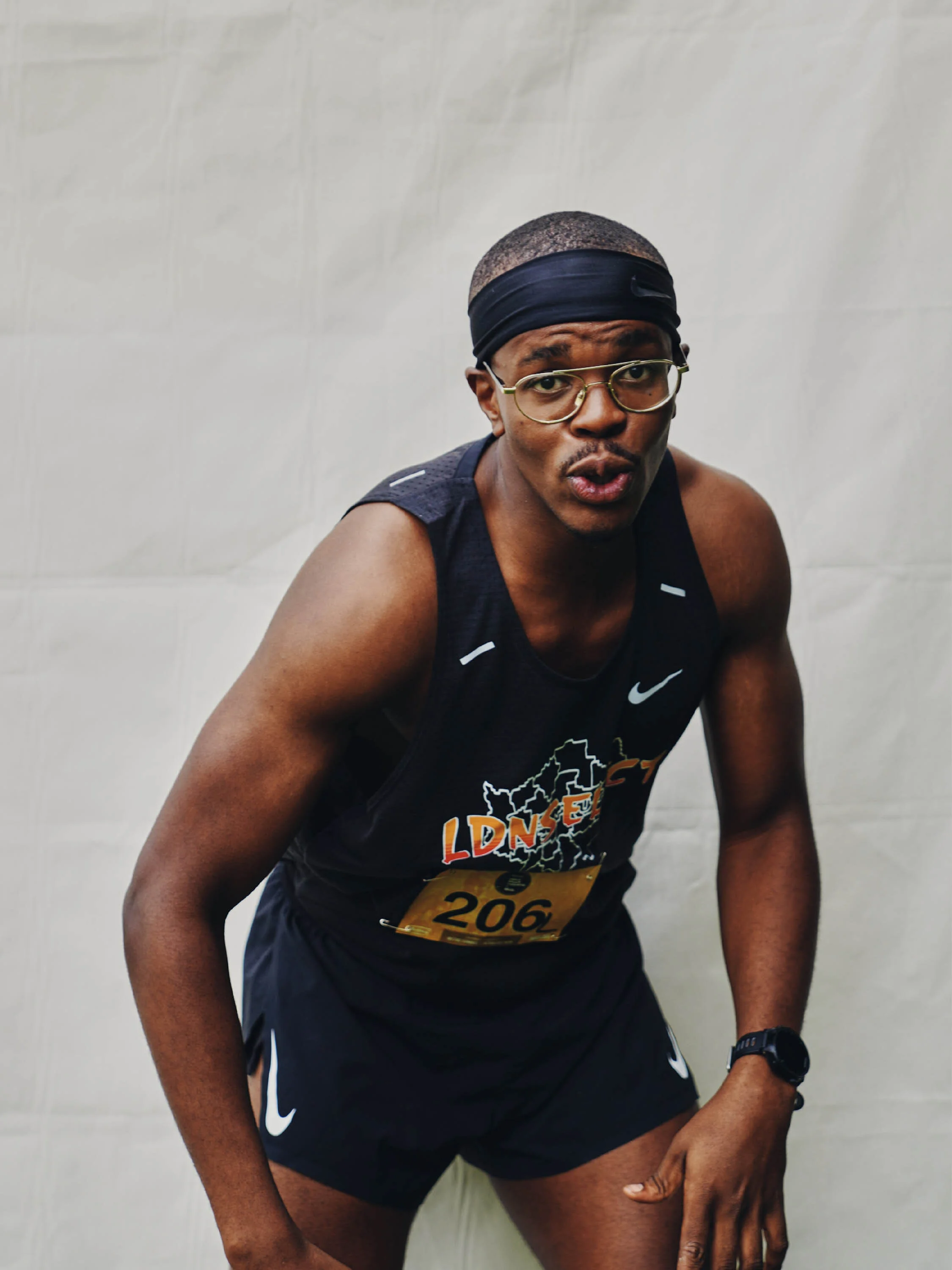 A male runner wearing glasses, a black headband, and athletic gear is leaning forward, appearing tired after a race.